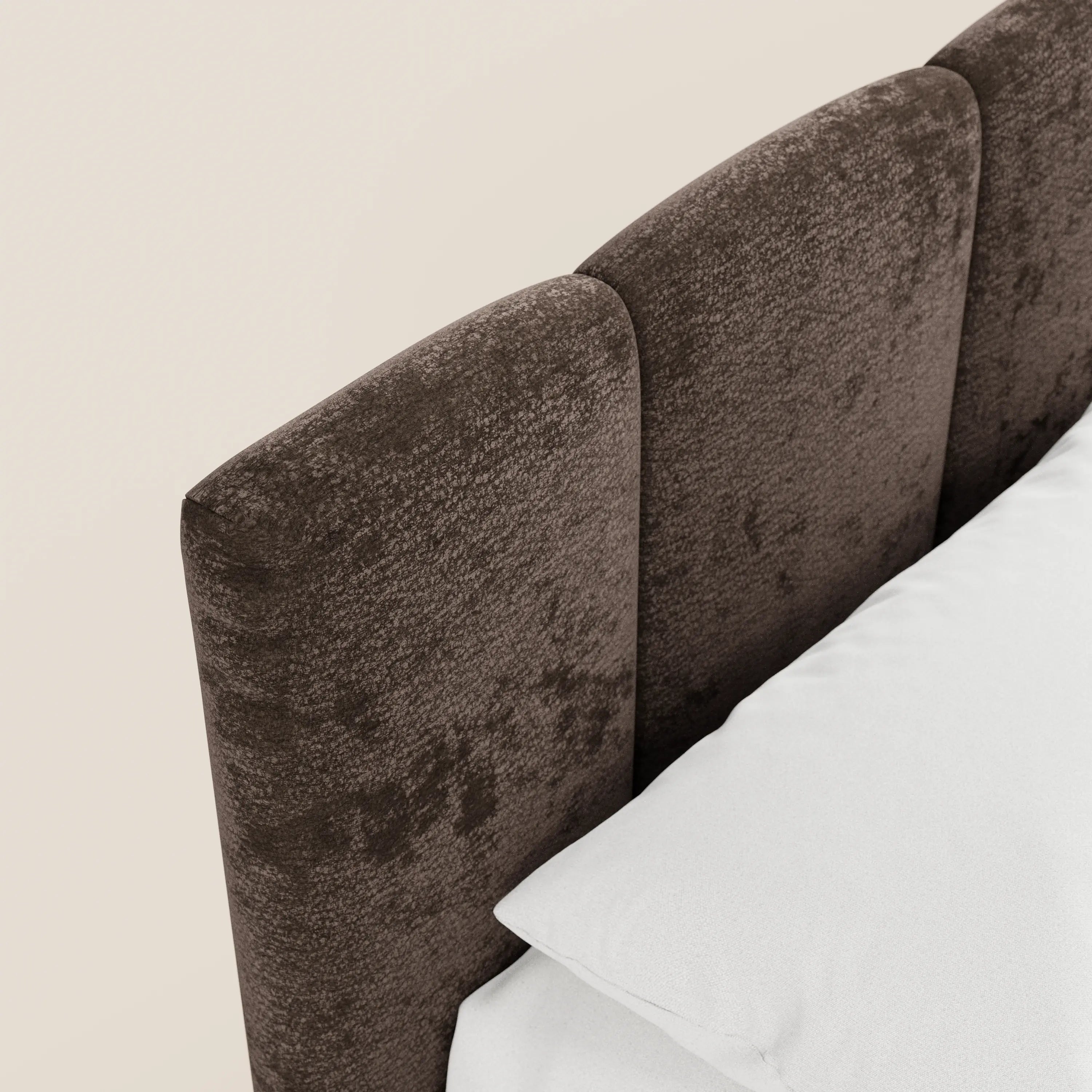 Una ripresa ravvicinata e obliqua del letto imbottito ARKAM con contenitore e testiera a lavorazione verticale di Letti.store mette in risalto il suo tessuto Oeko-Tex® marrone scuro e testurizzato e il suo design shabby chic, in un letto ben rifatto con biancheria da letto bianca su uno sfondo beige chiaro.
