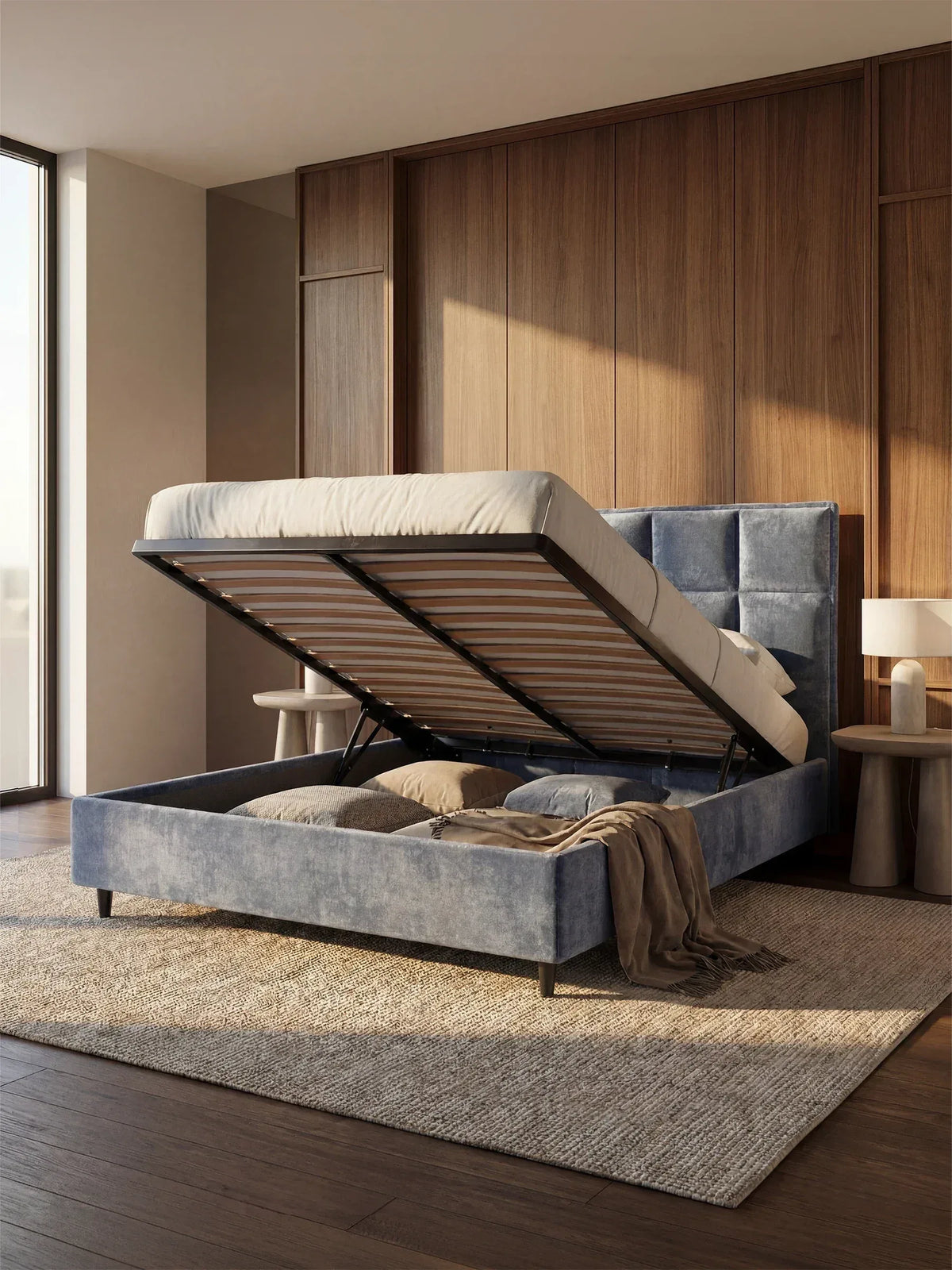 Una camera da letto moderna caratterizzata da un'ampia finestra, pareti rivestite in legno e dal letto imbottito AURO con contenitore e testiera capitonné alta in ciniglia Oeko-Tex® di Letti.store, raffigurato con la base imbottita aperta per mostrare l'ampio spazio di stivaggio sotto il letto. La biancheria da letto dai toni neutri e un morbido plaid completano il letto, mentre due comodini rotondi e un tappeto beige completano l’arredamento accogliente.