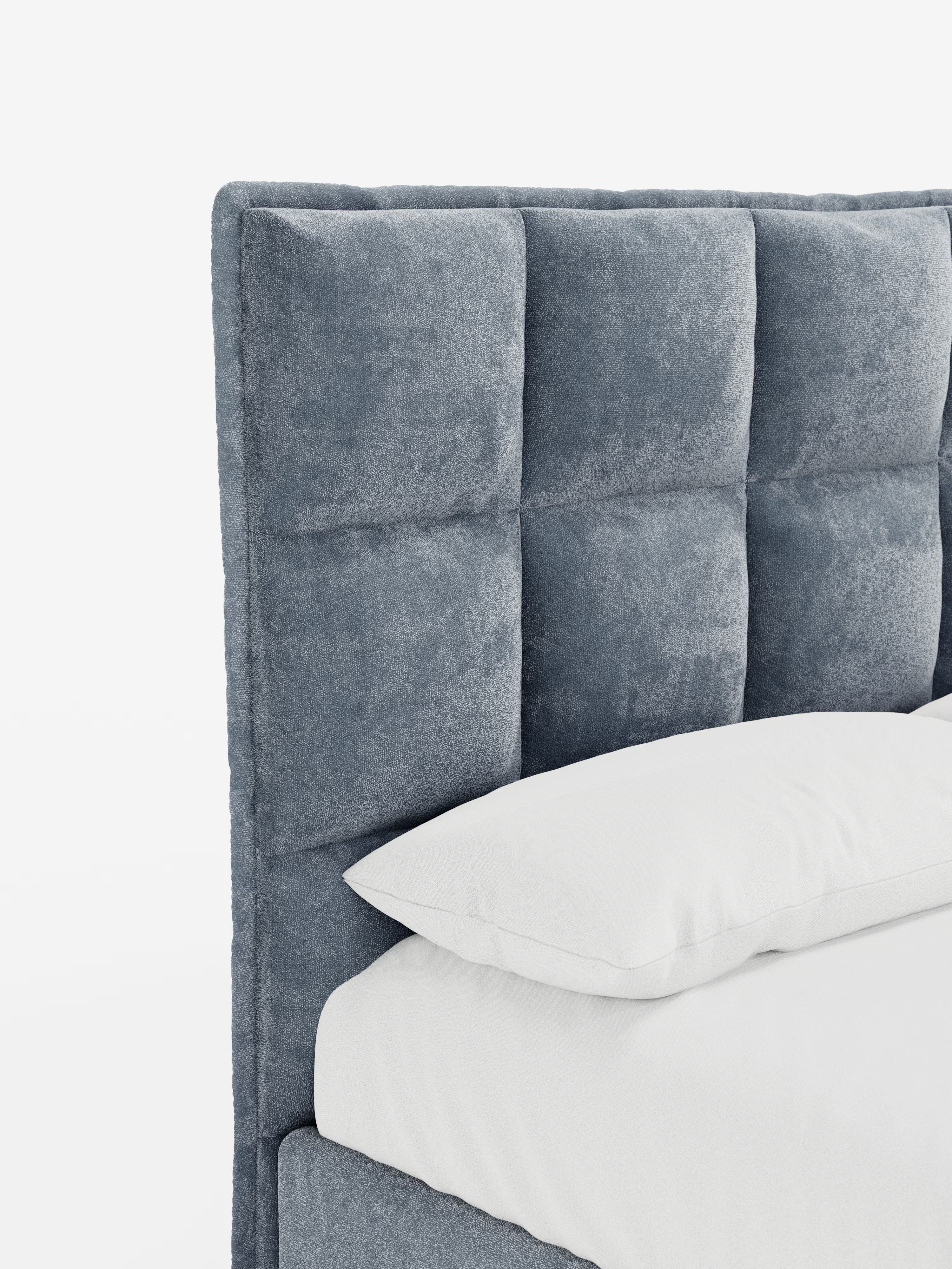 Un primo piano del letto imbottito con contenitore AURO di Letti.store mette in risalto la sua alta testiera capitonné, rivestita in morbido e soffice tessuto ciniglia grigio certificato Oeko-Tex®. Le sezioni quadrate trapuntate della testiera offrono una rivisitazione in chiave moderna del design classico. Un cuscino bianco poggia sul materasso, in perfetta armonia con la tonalità grigia della testiera. Lo sfondo è bianco a tinta unita.