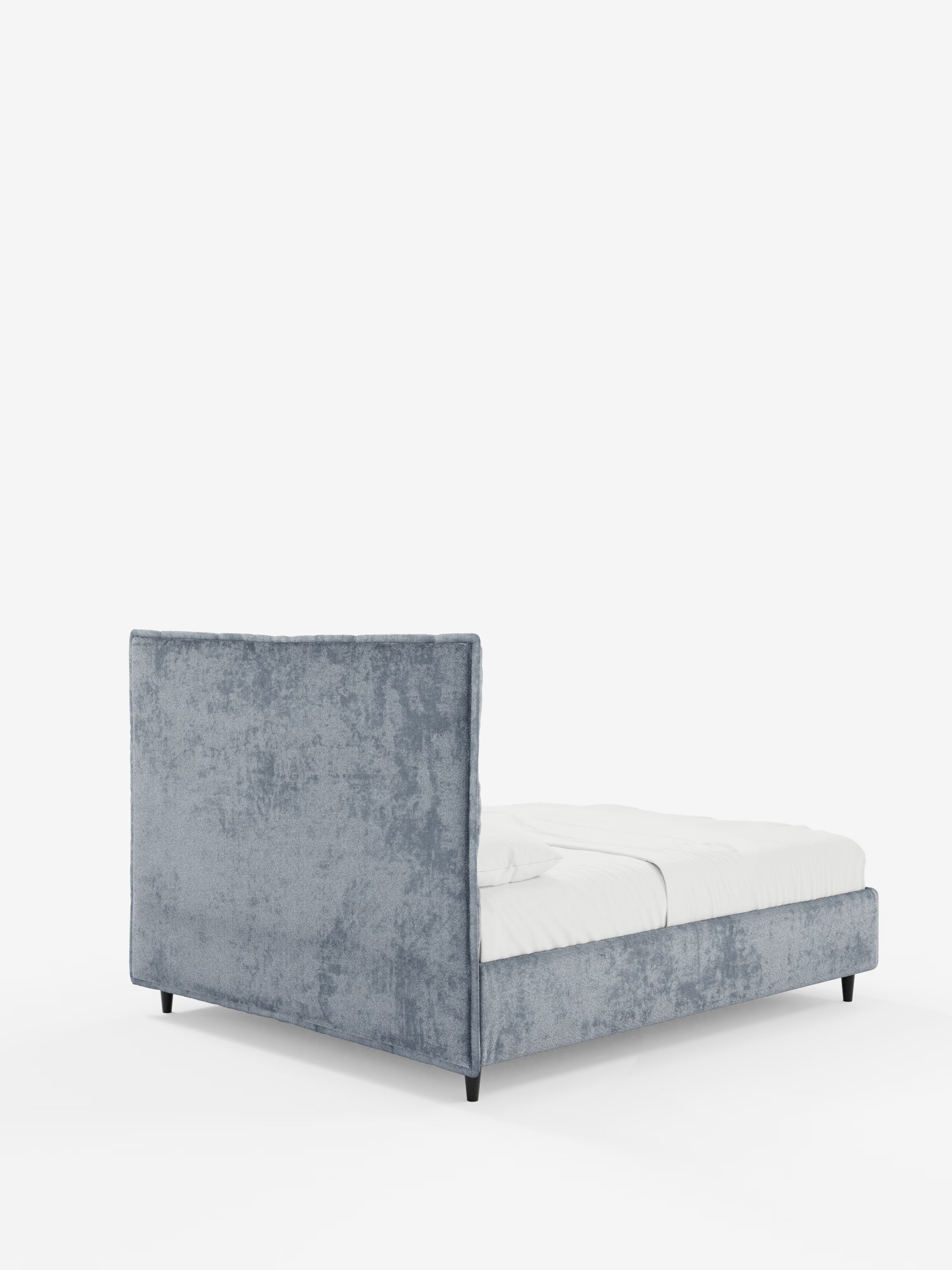Il letto imbottito AURO con contenitore di Letti.store presenta un rivestimento in ciniglia azzurra certificata Oeko-Tex® e una testiera alta e rettangolare con lavorazione capitonné. Il letto ha una pediera bassa, gambe corte nere ed è rivestito con biancheria da letto bianca. Fotografato su uno sfondo bianco a tinta unita, l’immagine mostra il letto da un’angolazione che ne mette in risalto sia la parte posteriore che quella laterale.