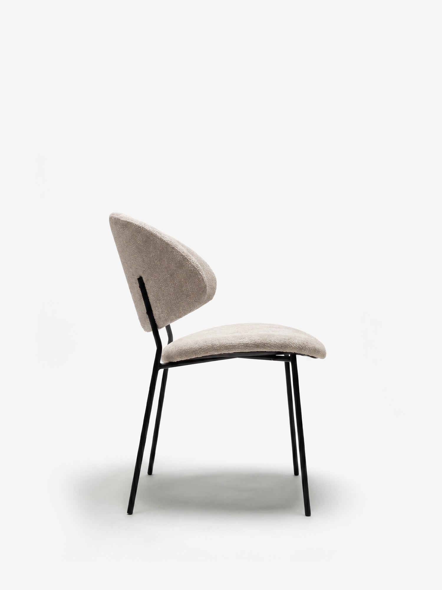 La sedia Bruma di Divani.Store è caratterizzata da un elegante tessuto goffrato, gambe in metallo nero inclinate e uno schienale avvolgente dal design minimalista; nella foto è raffigurata di profilo su uno sfondo bianco.