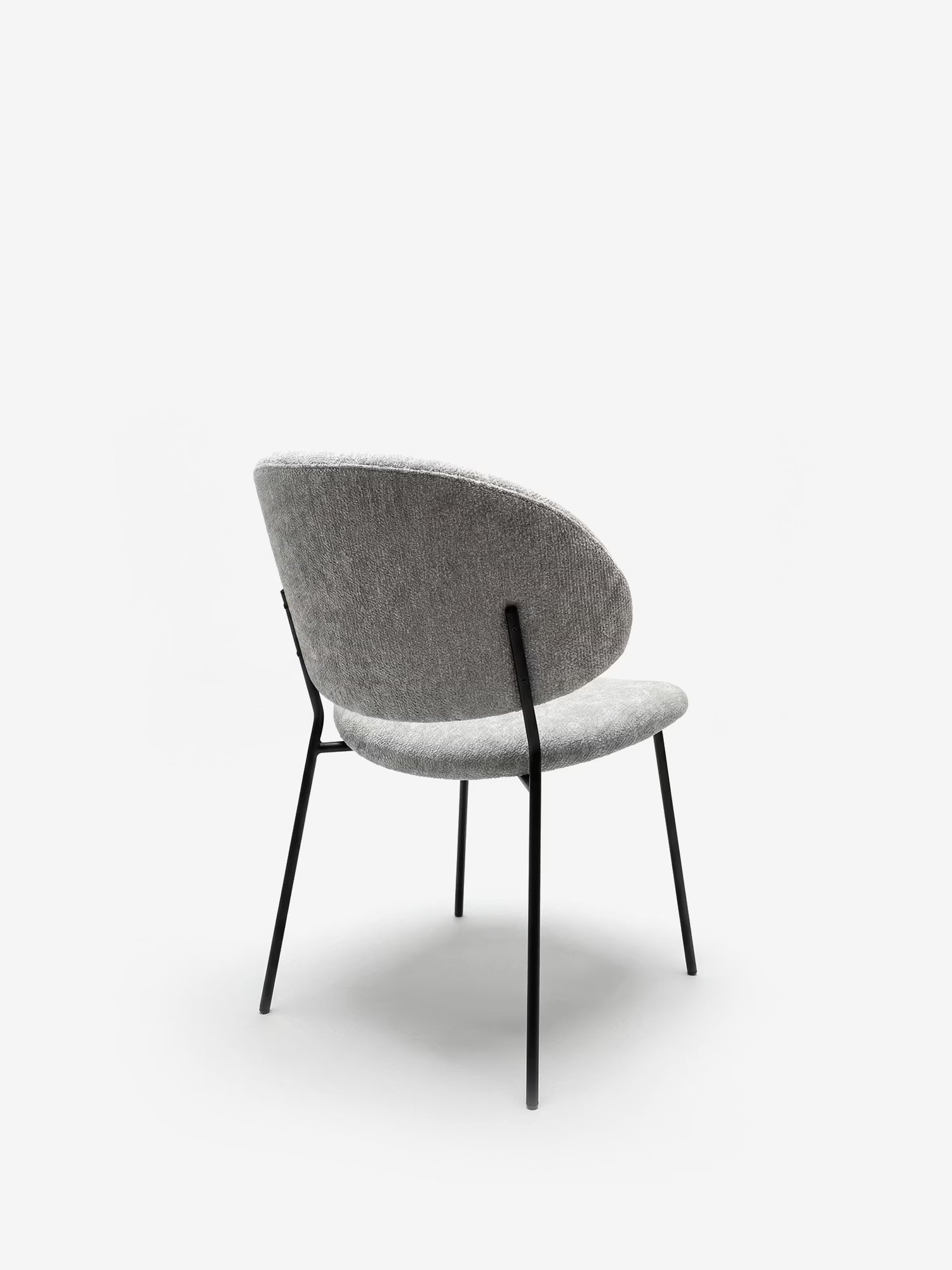 La Bruma di Divani.Store è una sedia dal design minimalista con schienale avvolgente in tessuto goffrato grigio e gambe sottili in metallo nero, fotografata di tre quarti su sfondo bianco.