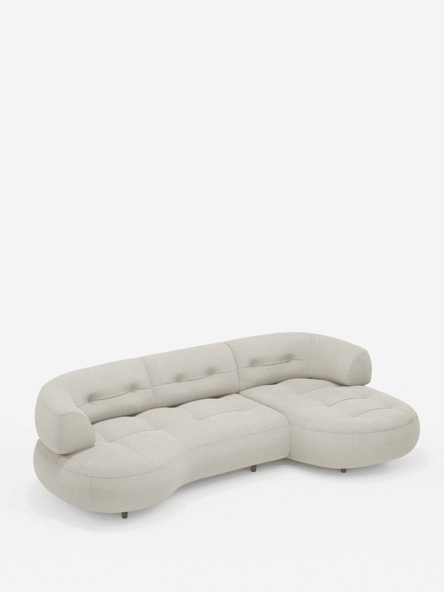 Un divano angolare Croissant color beige chiaro, dalla forma a fagiolo e con schienali scorrevoli, rivestito in morbido tessuto effetto cotone lavabile, modello T33 con cuscini trapuntati e schienali bassi, è posizionato su uno sfondo bianco.