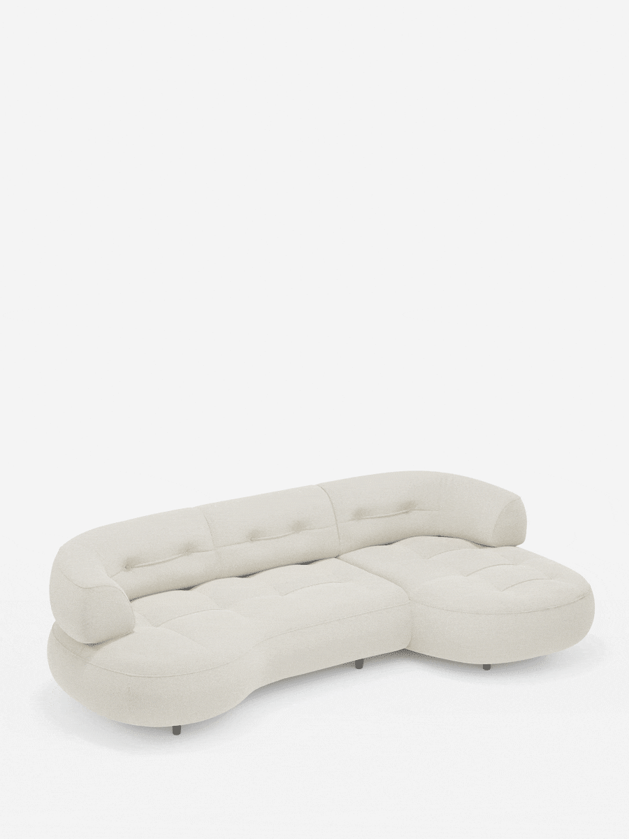 Un divano angolare Croissant color crema, dal design a forma di fagiolo e con schienali scorrevoli in morbido tessuto effetto cotone smacchiabile T33, caratterizzato da uno schienale curvo trapuntato con bottoni e da gambe scure dal profilo basso su sfondo bianco.