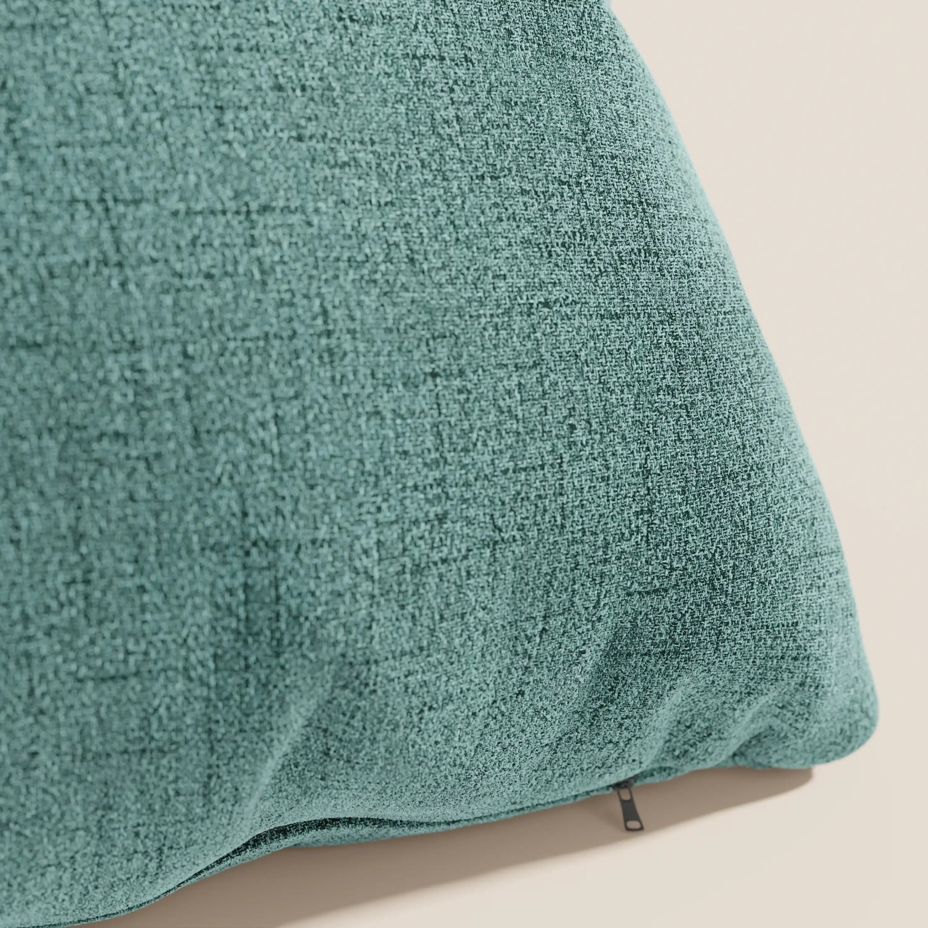 Un primo piano del cuscino Felis 45x45 cm di Divani.Store, in tessuto morbido impermeabile verde acqua T03, caratterizzato da una sottile trama a griglia e dalla linguetta nera della cerniera per il rivestimento sfoderabile su uno sfondo beige chiaro.