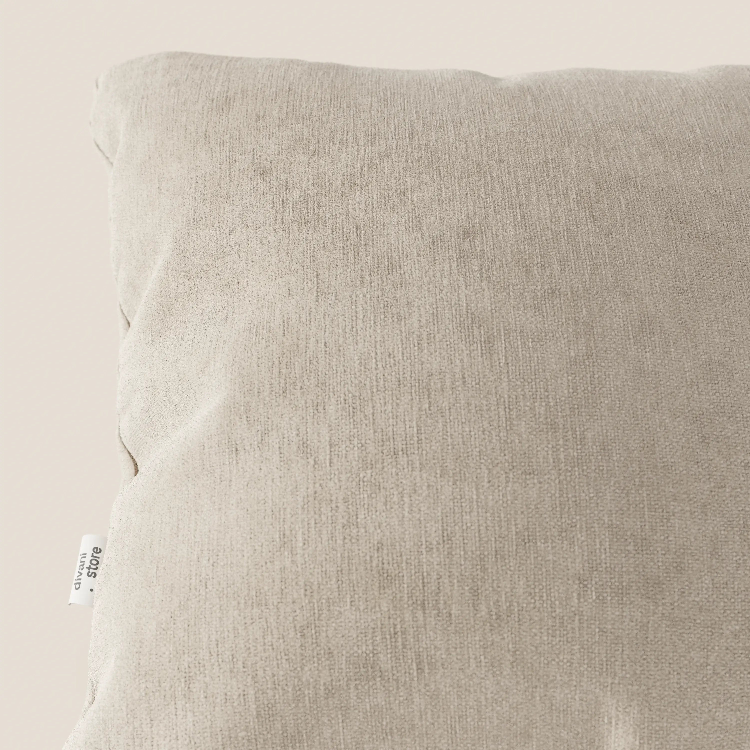 Un primo piano del Bolero cuscino di Divani.Store in beige chiaro mostra il suo tessuto T31 morbido, testurizzato, smacchiabile, impermeabile e irrestringibile. Una targhetta bianca con testo nero è visibile sulla cucitura sinistra su uno sfondo a tinta unita.