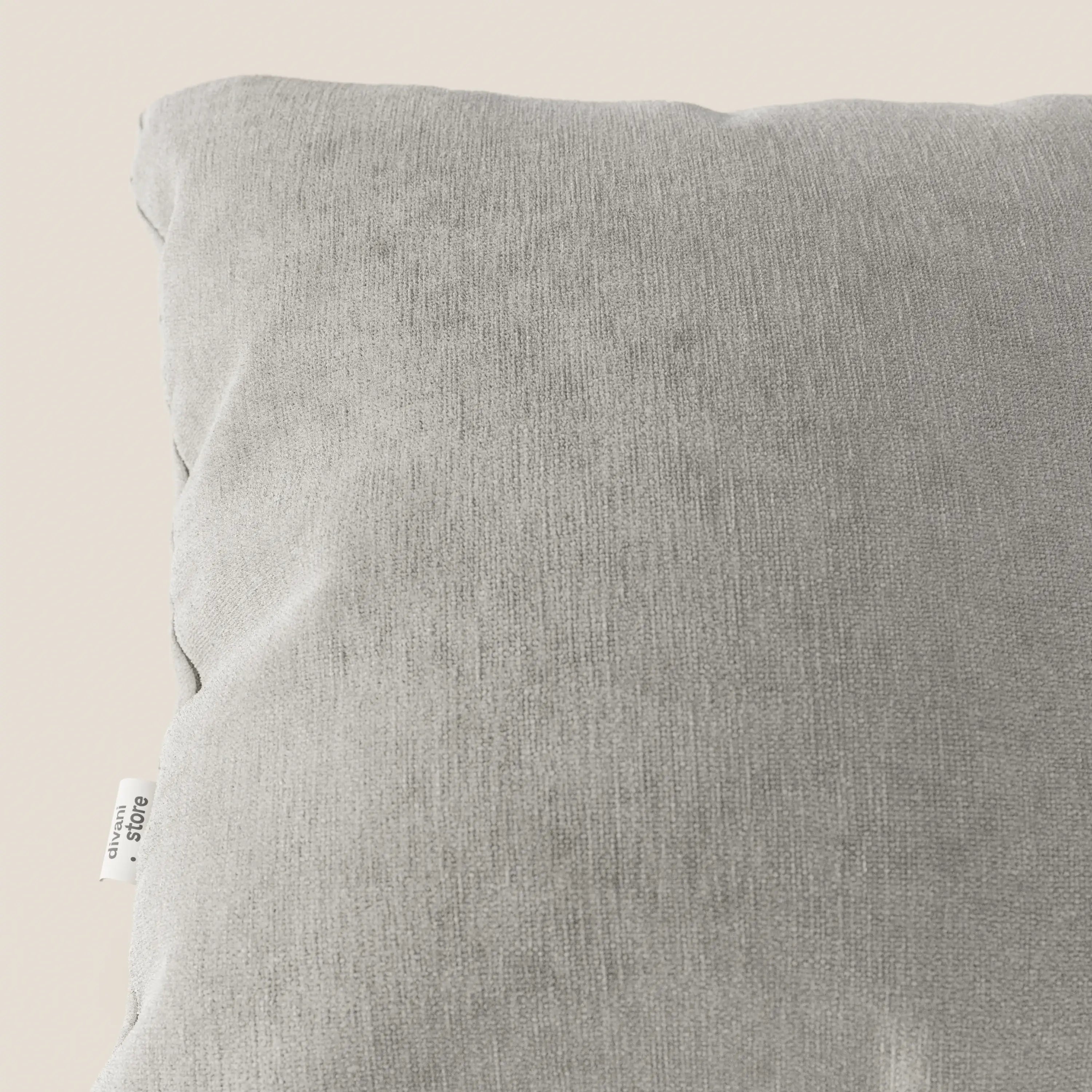Primo piano del cuscino Bolero di Divani.Store in grigio chiaro, realizzato in morbido tessuto T31 antimacchia, impermeabile e non restringibile, su sfondo beige con una piccola etichetta bianca sulla cucitura in basso a sinistra.