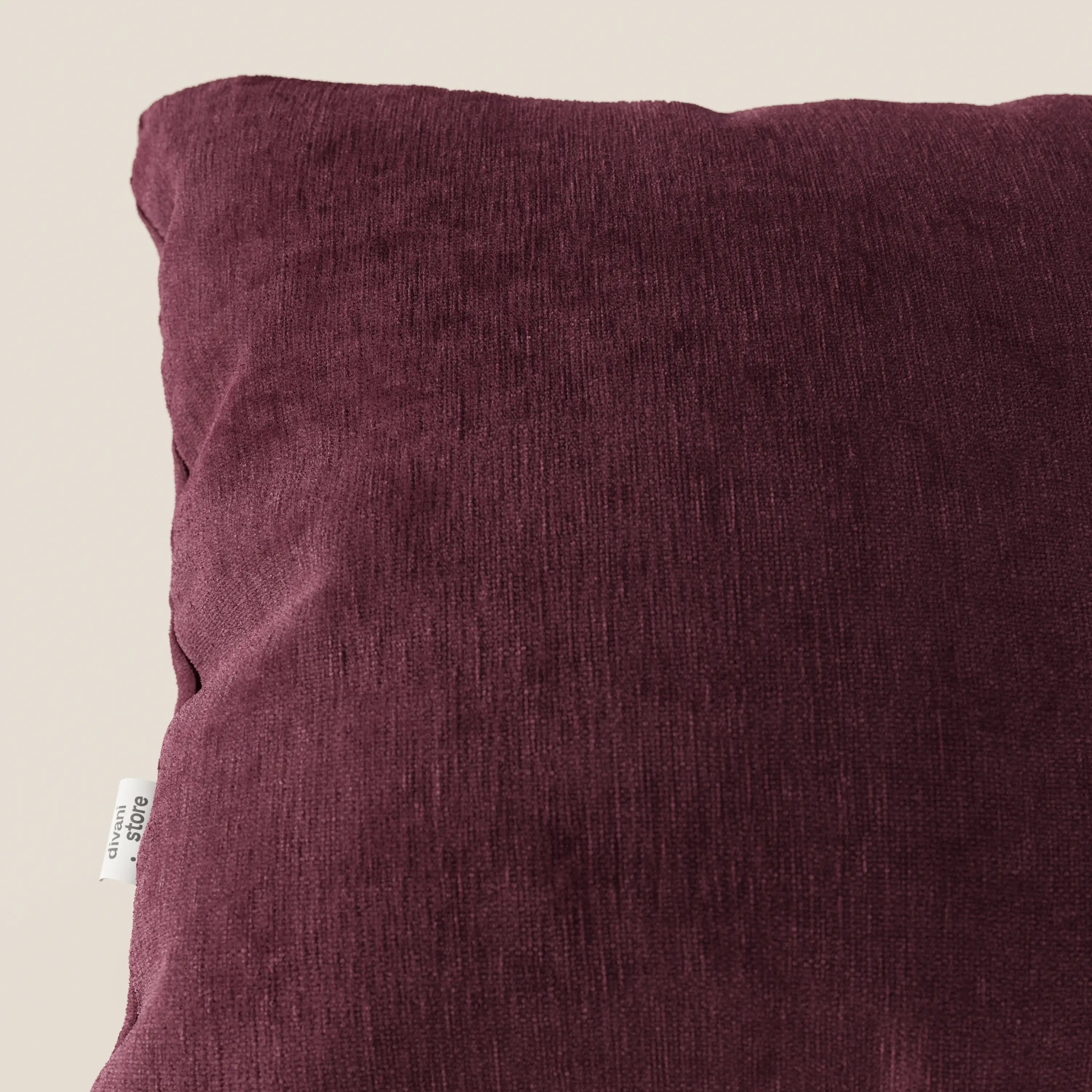 Primo piano del cuscino Bolero di Divani.Store, un cuscino quadrato in morbido tessuto T31 antimacchia, impermeabile e antirestringimento. Il cuscino, di colore bordeaux scuro, presenta una piccola etichetta bianca sulla sinistra ed è inserito in uno sfondo beige semplice.