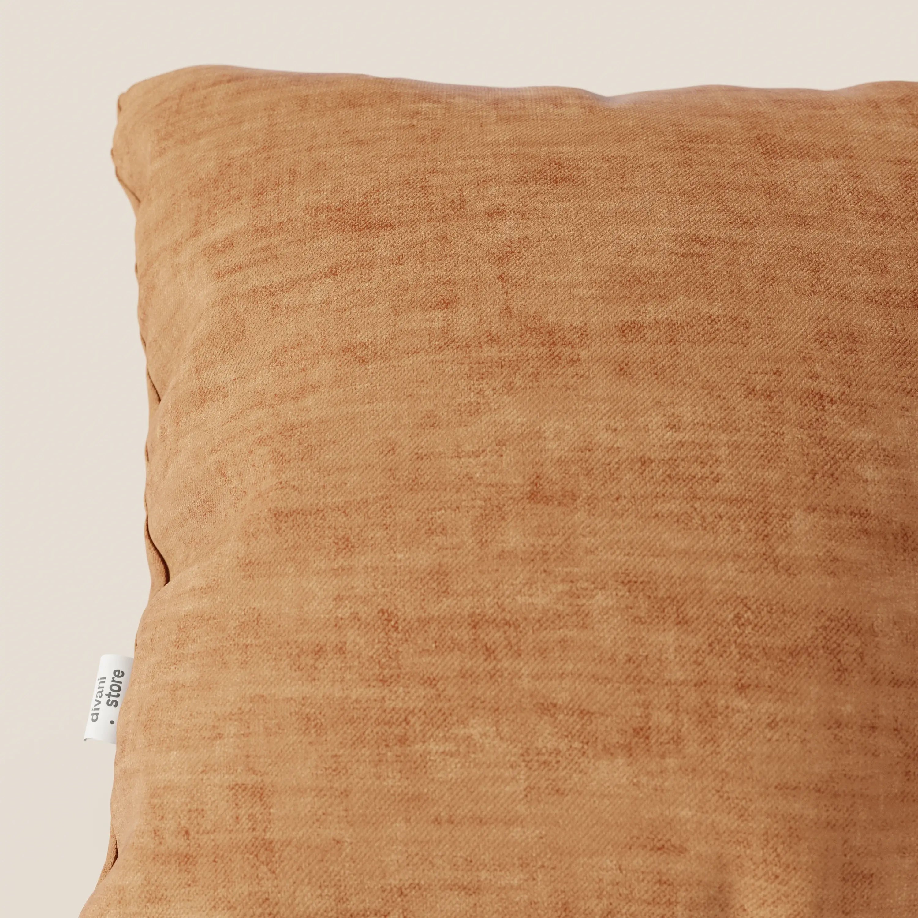Primo piano del cuscino Sofia di Divani.Store in velluto nuvolato T35 color ruggine con una texture sottile. Una targhetta in tessuto bianco con testo nero è cucita nella cucitura sinistra. Lo sfondo è un semplice beige chiaro.