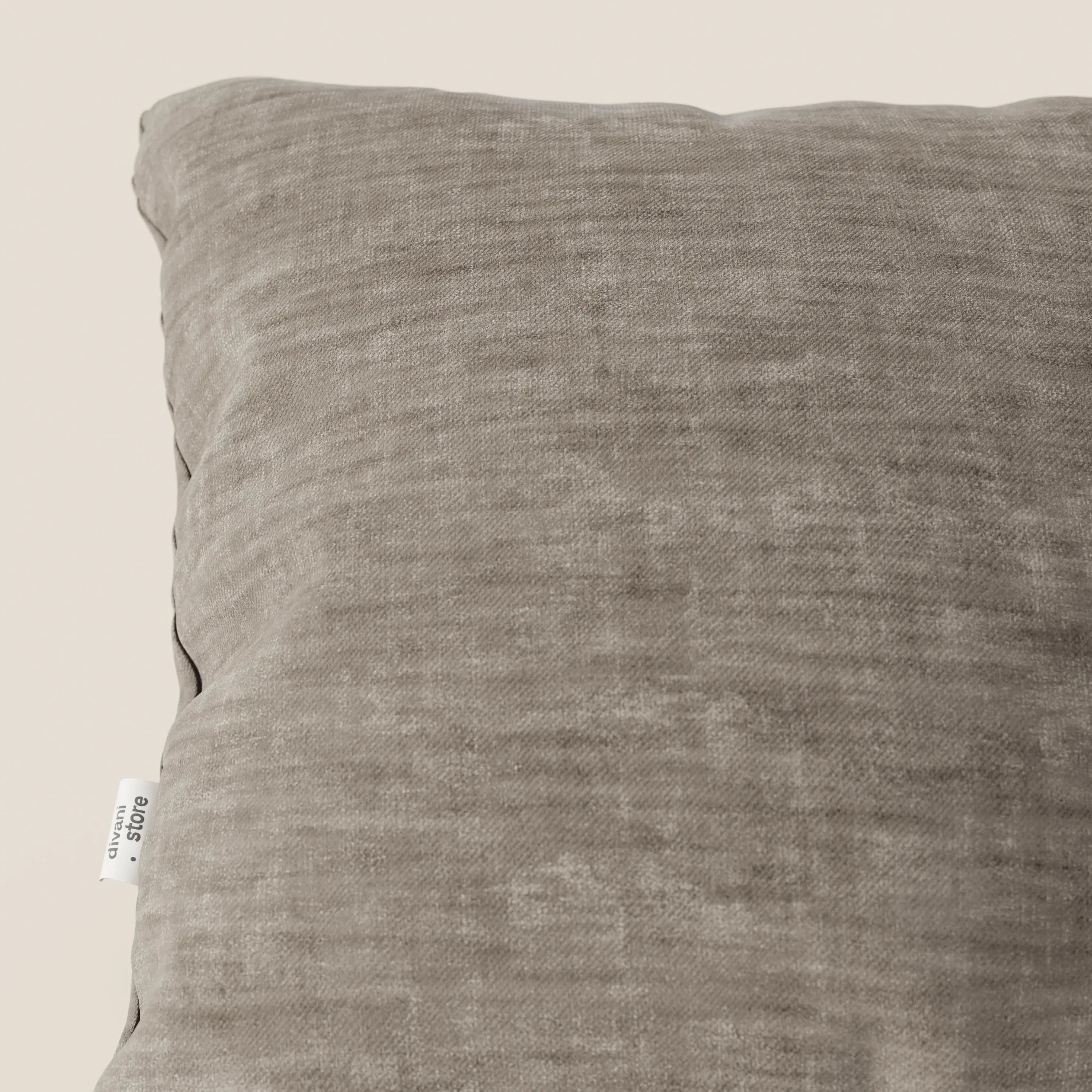 Primo piano del cuscino Sofia di Divani.Store in marrone chiaro, caratterizzato da una trama a vista e da una piccola etichetta bianca sulla cucitura sinistra. Realizzato in morbido tessuto vellutato Easy Clean T35, antimacchia e impermeabile. Il fondo è in tinta unita e di colore chiaro.