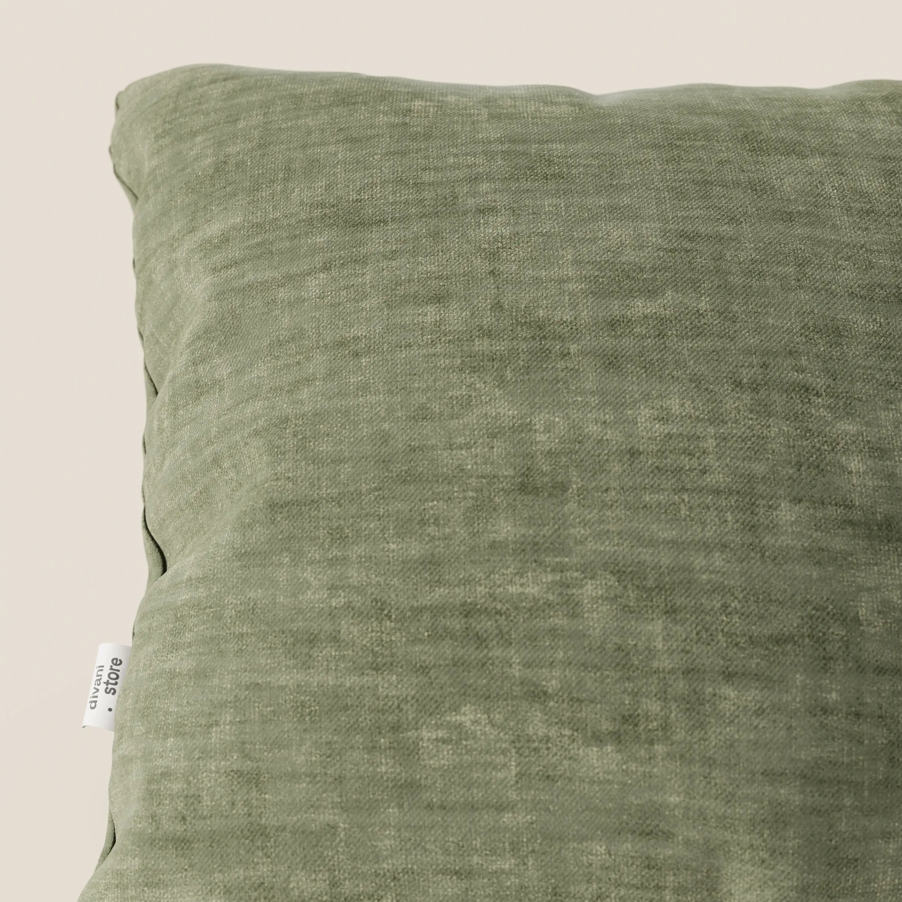 Primo piano del cuscino Sofia di Divani.Store, realizzato in morbido velluto verde nuvolato (Easy Clean T35), caratterizzato da una piccola etichetta bianca con testo nero sulla cucitura sinistra. Lo sfondo è bianco sporco e si vede solo una parte del cuscino.