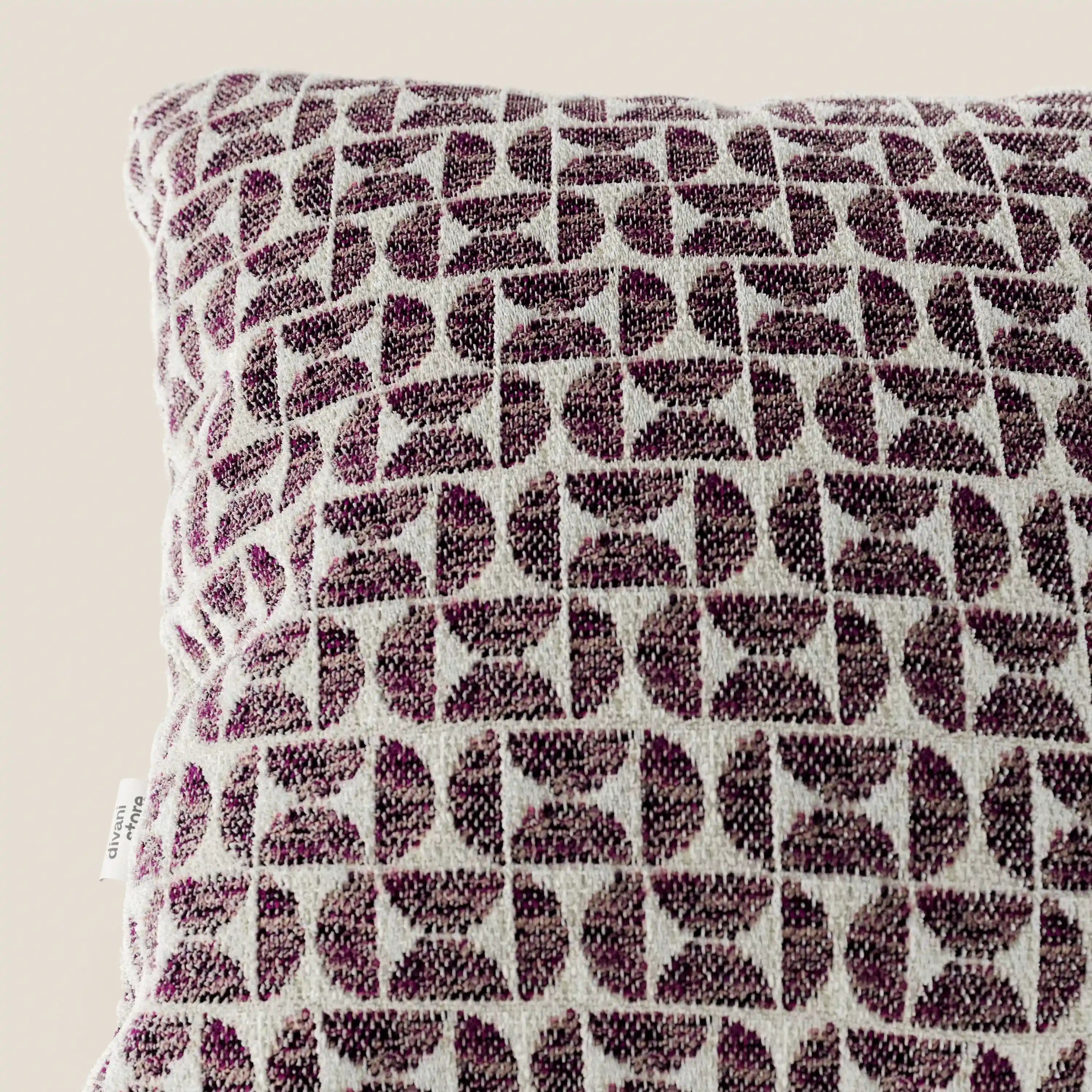 Un primo piano del cuscino Naxos Decoro 45x45 in lino di Divani.Store, che presenta un elegante motivo geometrico nei toni del viola e del bianco, realizzato in tessuto intrecciato con trama in rilievo e dotato di una piccola etichetta bianca sul bordo sinistro.
