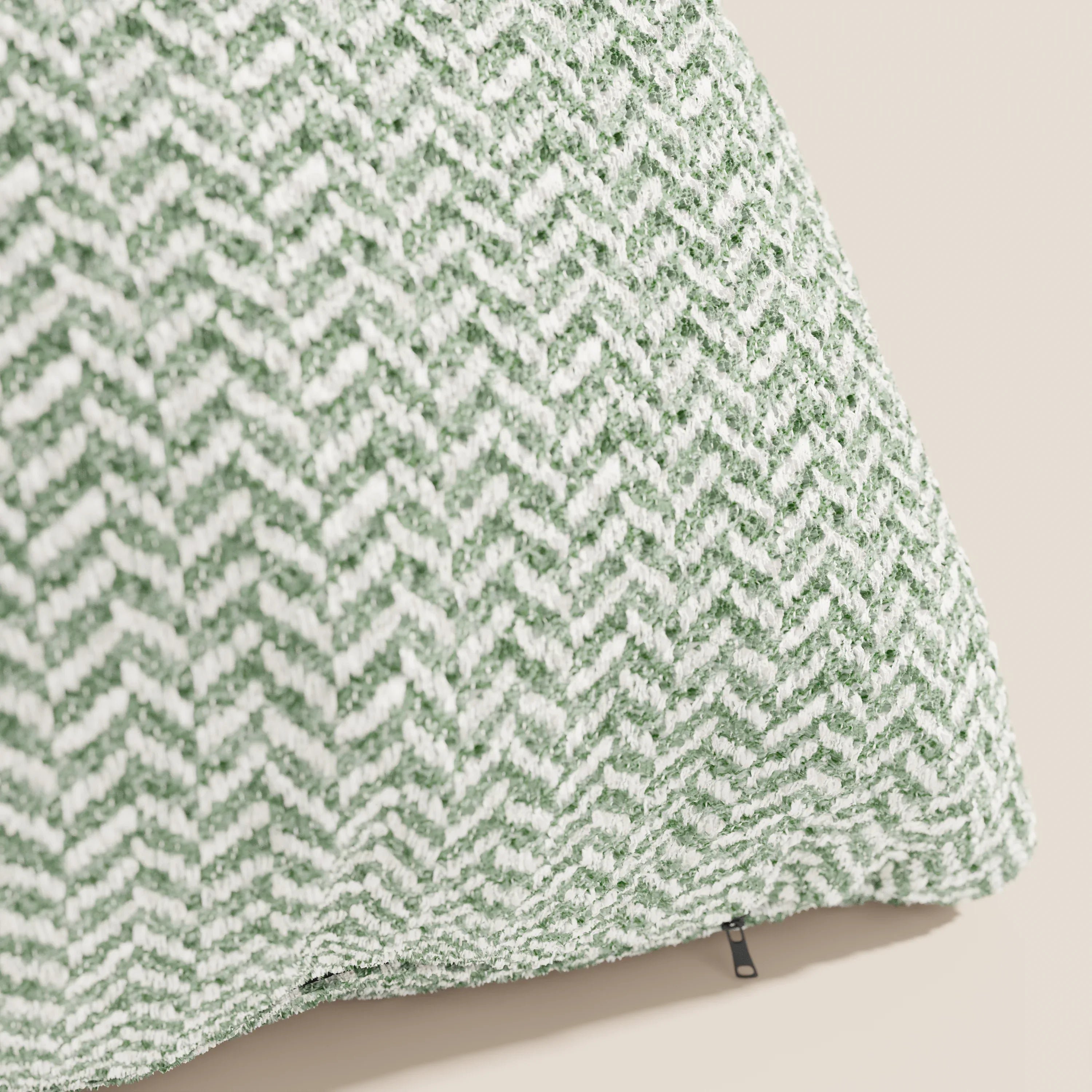 Un primo piano del cuscino Sorrento 45x45 in Lino di Divani.Store ne mette in risalto il motivo a spina di pesce verde e bianco su tessuto testurizzato, con una cerniera nera, il tutto su uno sfondo beige neutro.