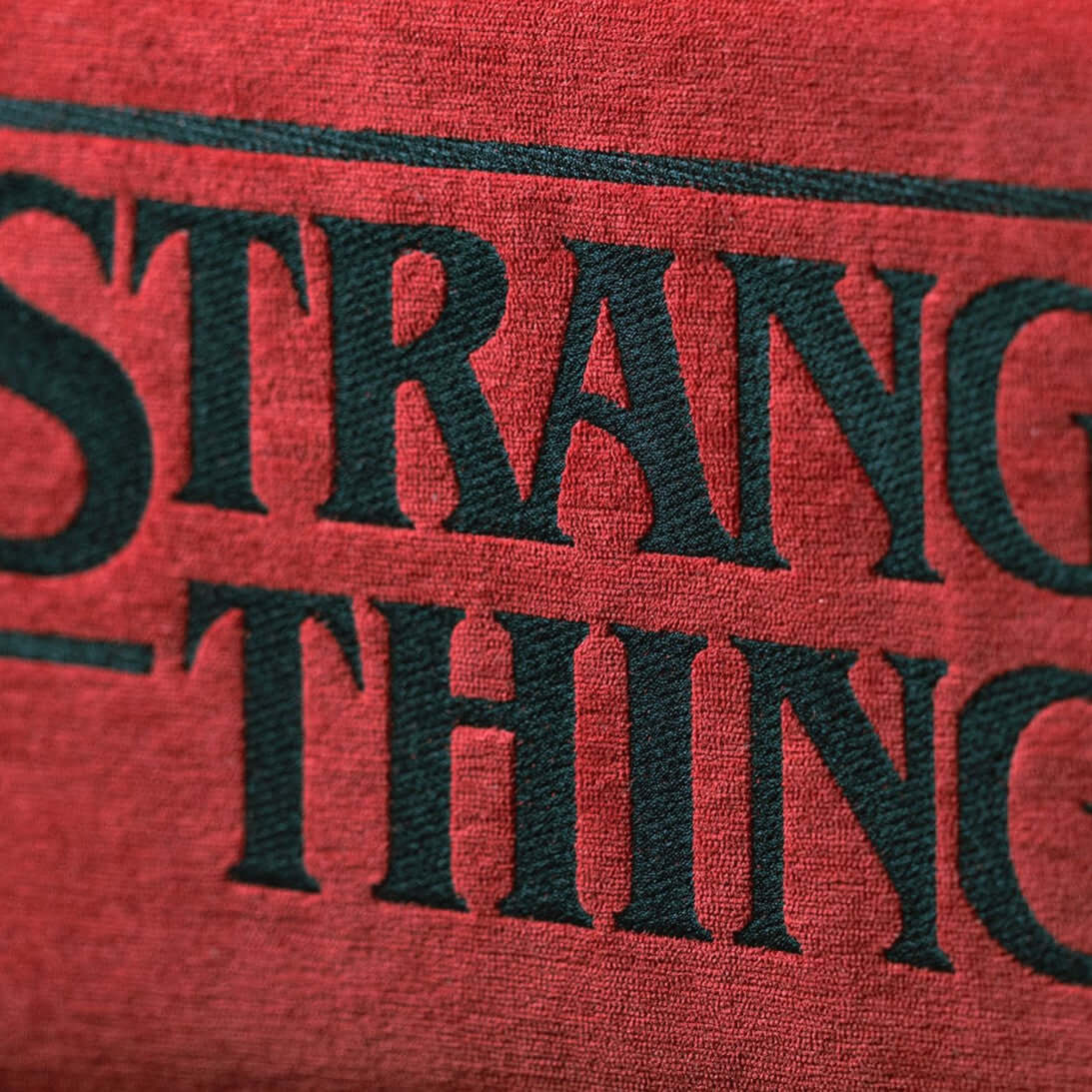Primo piano del tessuto rosso antimacchia del Cuscino Stranger Things Limited Edition con Ricamo (45x45) di Divani.Store, che mostra il testo serif nero in grassetto con "STRANG" e "THING" parzialmente visibili e la trama dettagliata del tessuto.