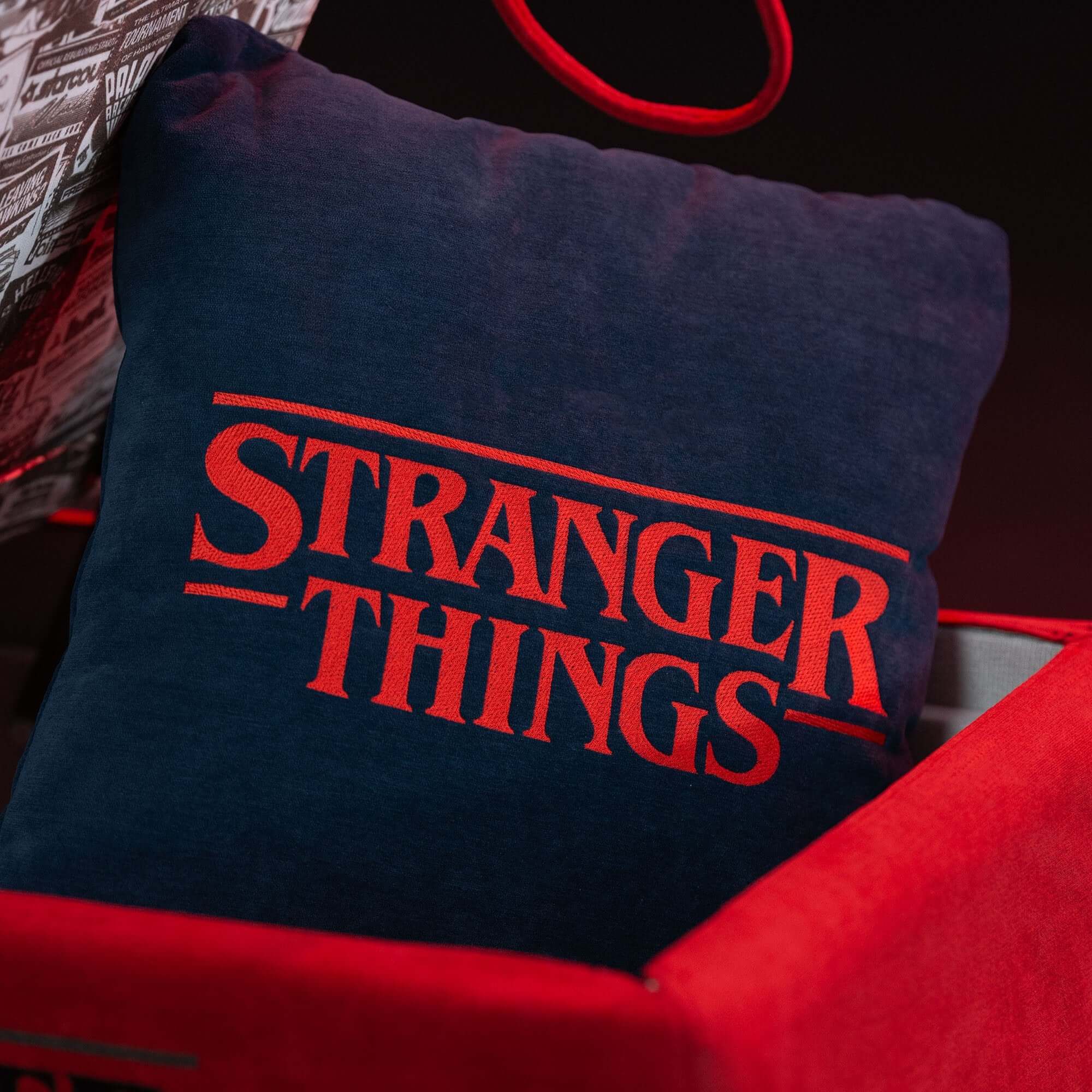 Il Cuscino Stranger Things Limited Edition con Ricamo di Divani.Store è caratterizzato da un logo rosso su tessuto blu antimacchia 45x45, presentato in una scatola rossa in un ambiente scuro ed elegante con tessuti a fantasia vicini.
