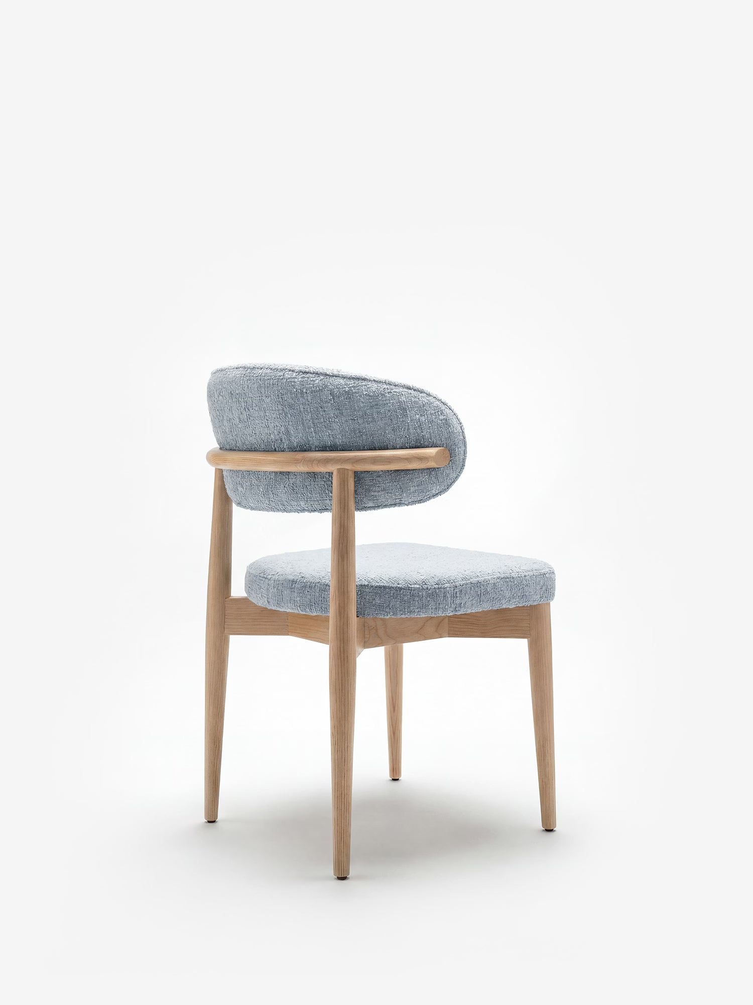 La sedia imbottita Dafne di Divani.Store presenta una struttura in rovere naturale massiccio con gambe in legno chiaro, uno schienale arrotondato e parzialmente ricurvo e un rivestimento in ciniglia azzurra certificato OEKO-TEX® — qui raffigurata su uno sfondo bianco a tinta unita.