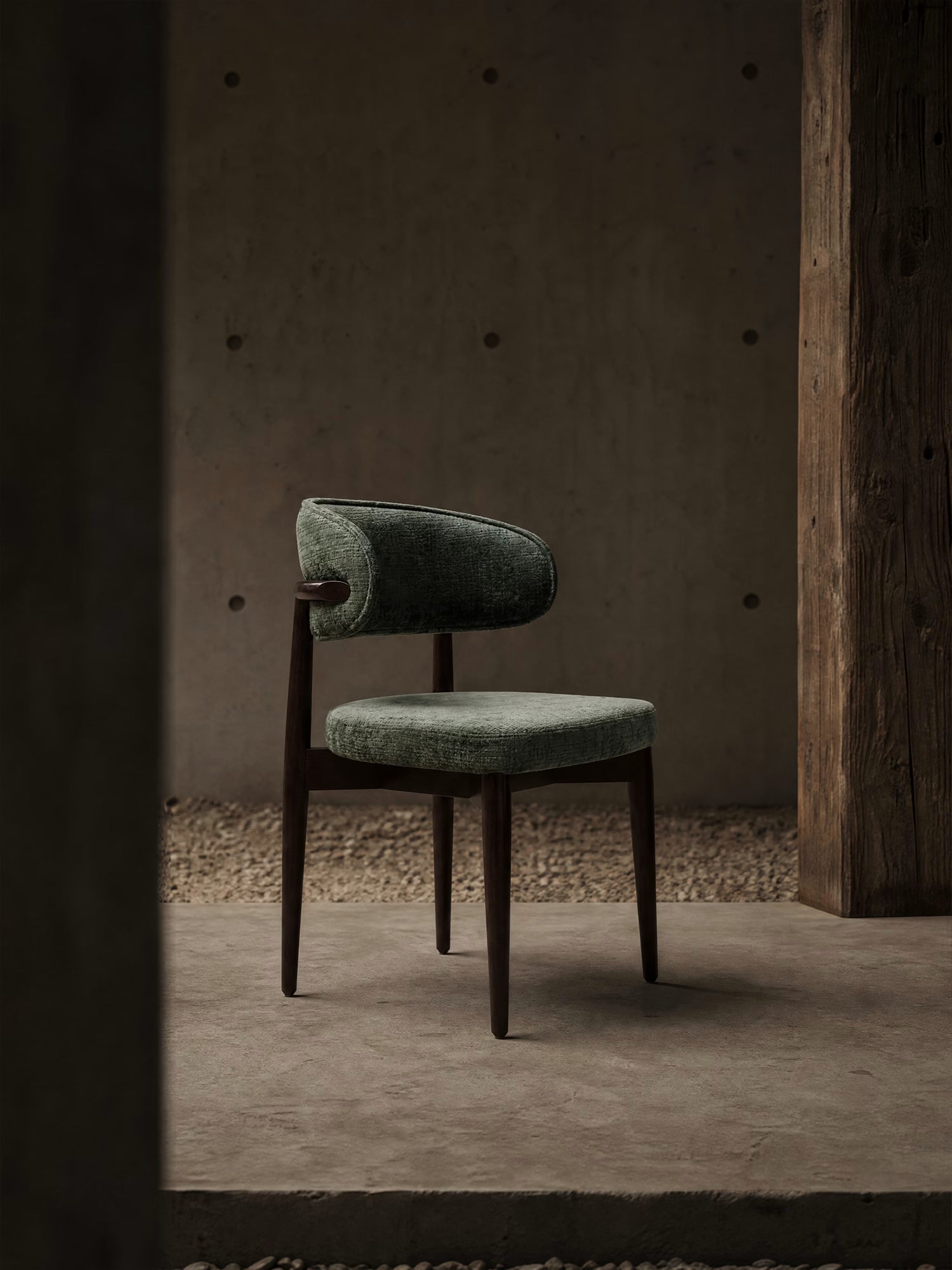 La sedia Dafne di Divani.Store presenta una seduta in ciniglia verde oliva certificata OEKO-TEX®, uno schienale curvo e una struttura in legno massello di wengé, conferendo eleganza e comfort a un ambiente minimalista con pareti in cemento e colonne in legno.