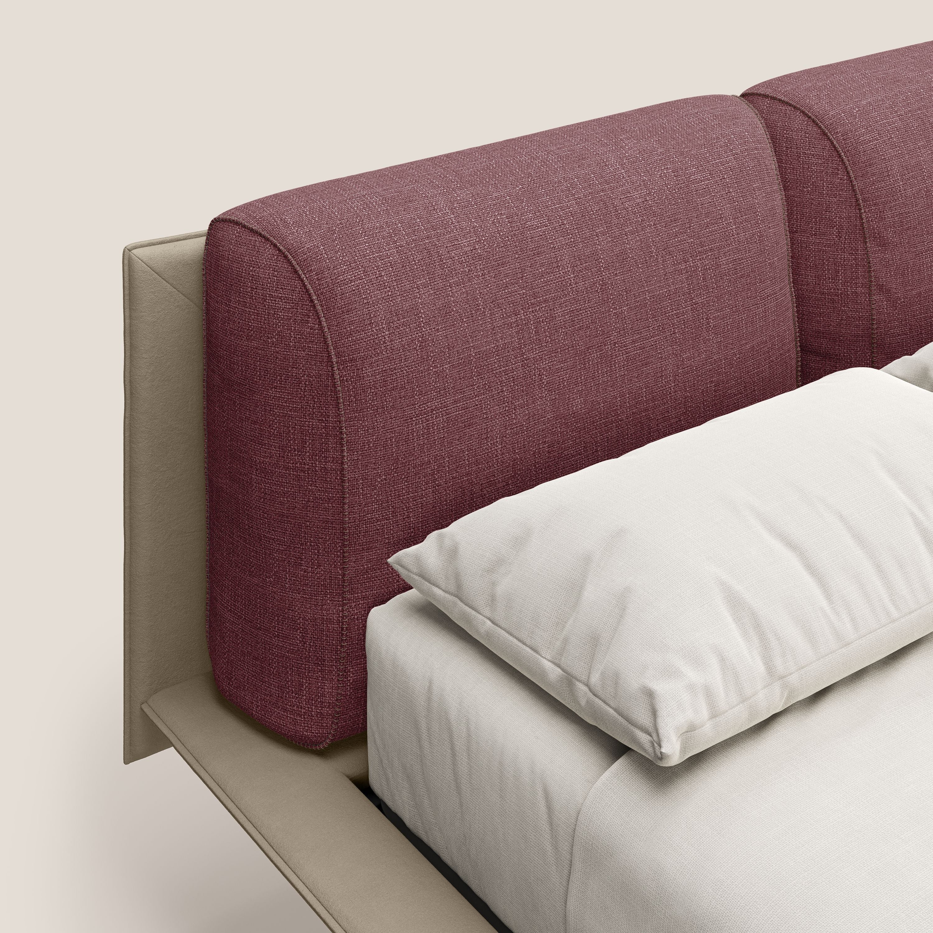 Primo piano del letto di design Petra di Letti.store, con testata in tessuto impermeabile bicolore T11 rosso e T06 tortora, struttura in morbido beige, cuscini in piuma e piedi in mogano. Il letto è confezionato in modo ordinato con biancheria da letto color crema su uno sfondo neutro.