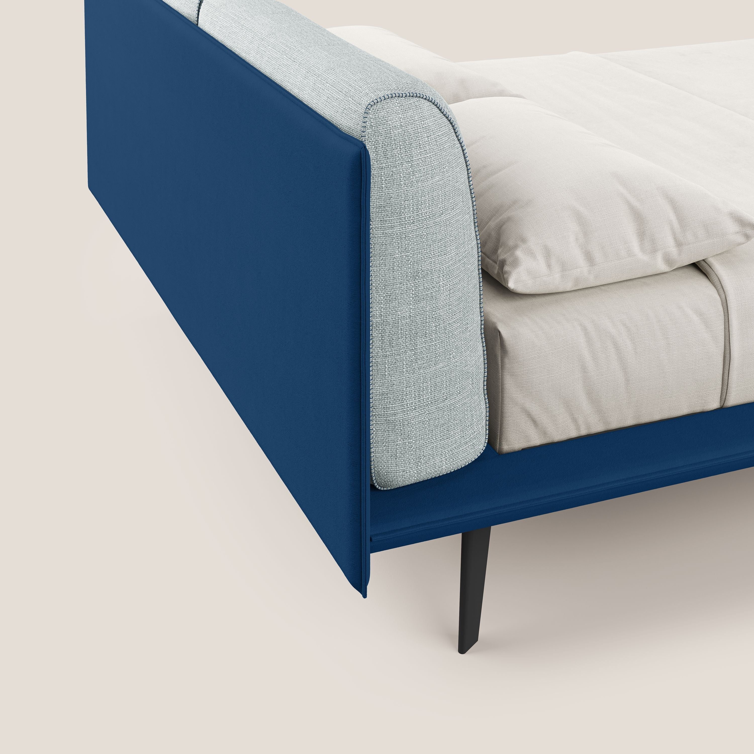 Primo piano del letto di design Petra di Letti.store, caratterizzato dal tessuto bicolore impermeabile T11 e T06 blu ghiaccio, dai cuscini in piuma, dalla biancheria da letto color crema e dalle sottili gambe in mogano. Lo sfondo neutro evidenzia il suo stile moderno e minimalista.