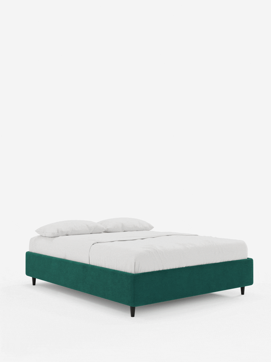 Un letto sommier ERON verde smeraldo, imbottito e dotato di vano contenitore in tessuto impermeabile certificato Oeko-Tex, con gambe affusolate scure, rivestito con lenzuola bianche immacolate e due cuscini su sfondo bianco.