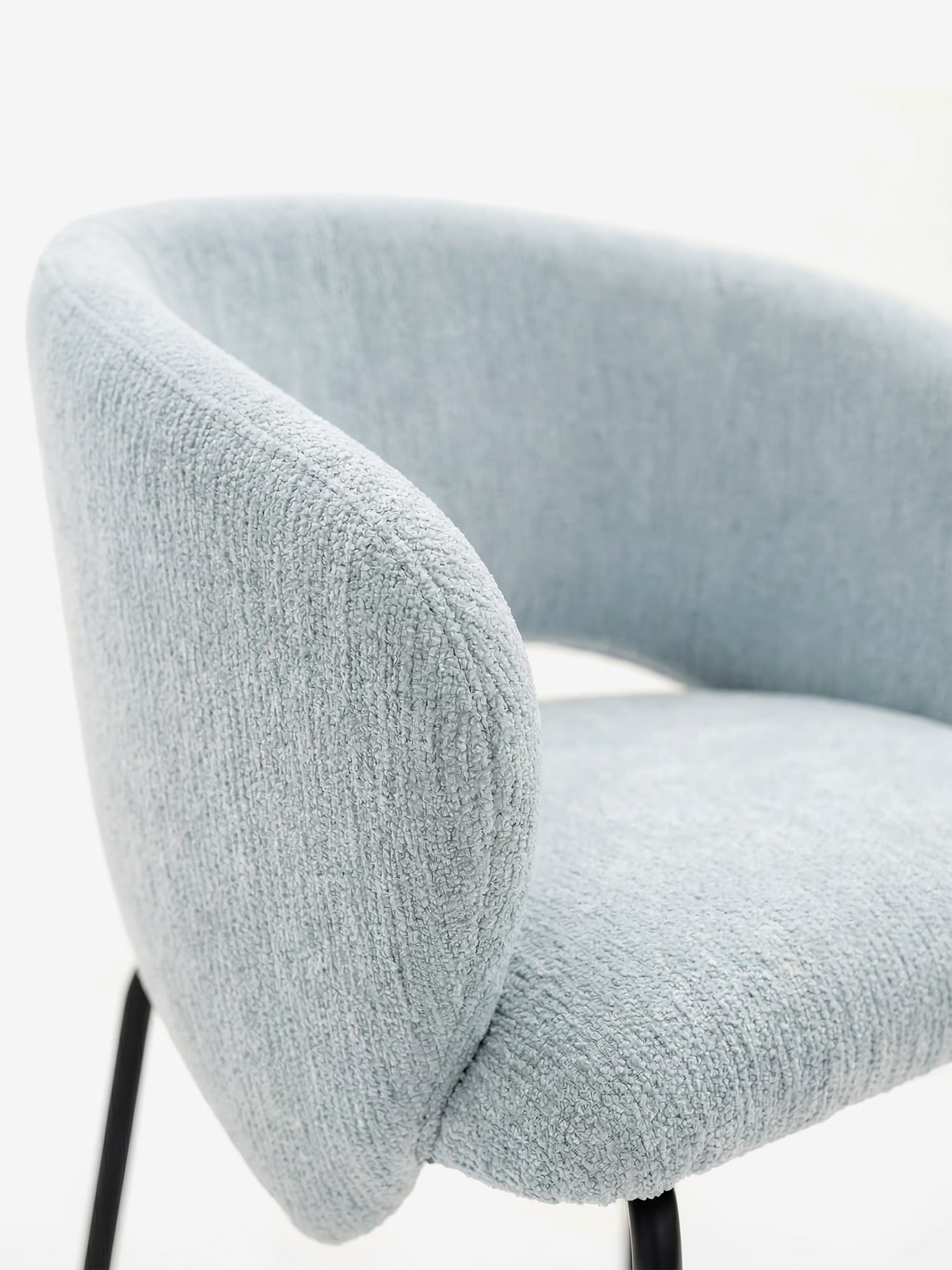 Un primo piano della sedia imbottita Fosca di Divani.Store, caratterizzata da braccioli di design, rivestimento in morbido tessuto azzurro certificato OEKO-TEX® e sottili gambe in metallo nero, su uno sfondo bianco a tinta unita.