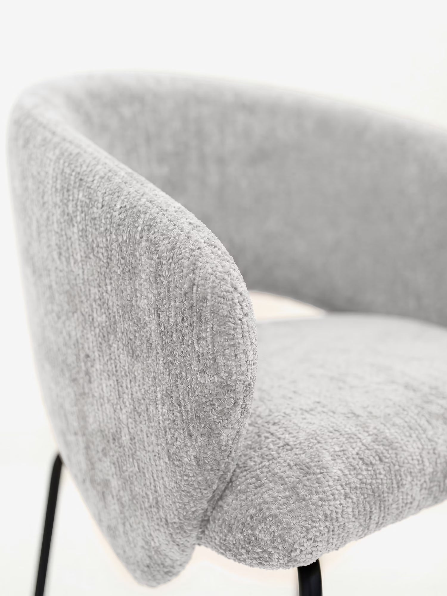 Primo piano della sedia imbottita Fosca di Divani.Store, caratterizzata da uno schienale curvo, braccioli, rivestimento in morbido tessuto certificato OEKO-TEX® color grigio chiaro e sottili gambe in metallo nero, su uno sfondo bianco a tinta unita.