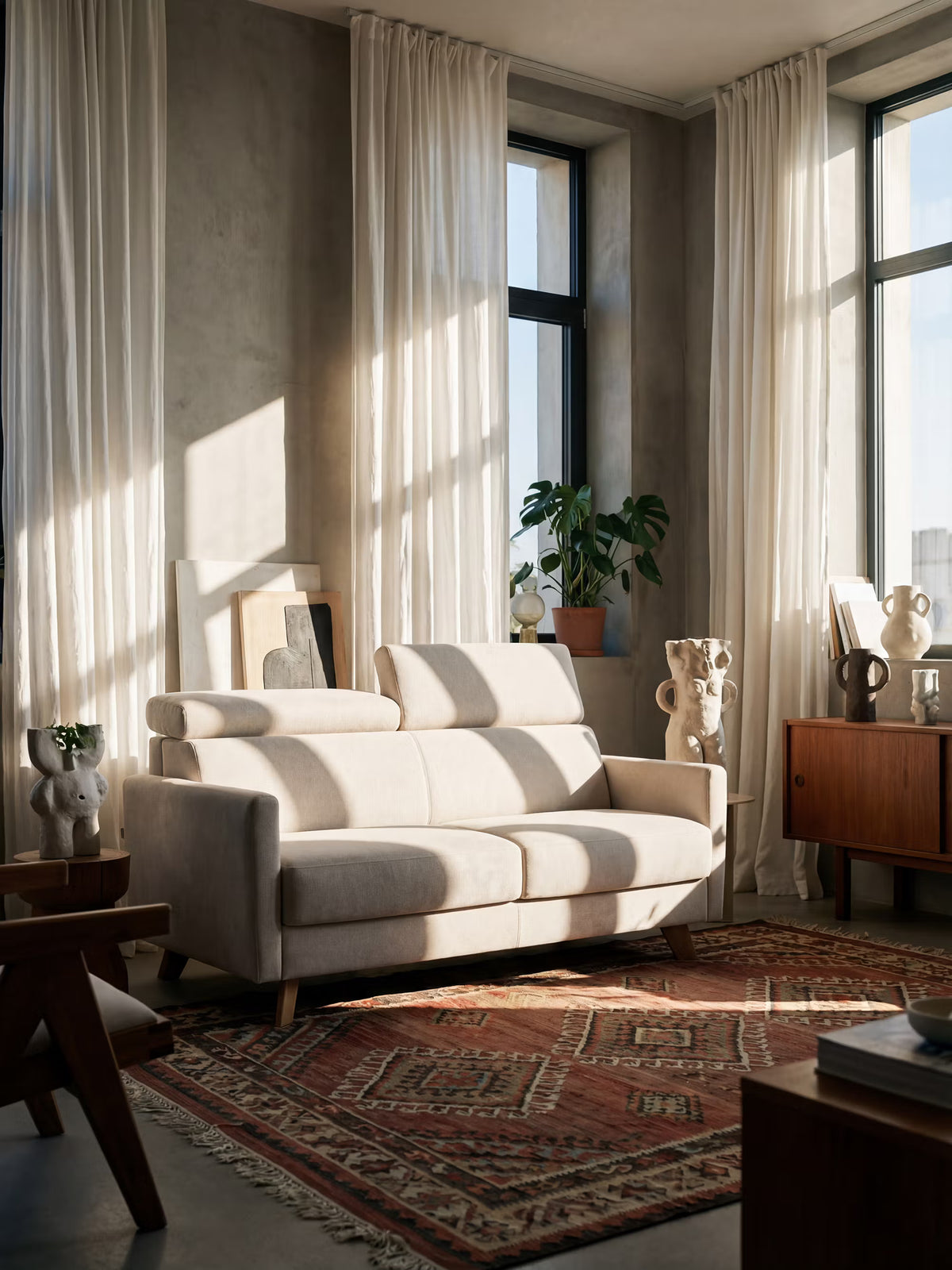Un soggiorno inondato di luce ospita il divano letto Francy con poggiatesta regolabili di Divani.Store, sfoderabile al 100% in elegante tessuto smacchiabile T30, posizionato su un tappeto a motivi rossi. Lo spazio è circondato da mobili in legno, elementi decorativi scultorei, una pianta appoggiata su una credenza e ampie finestre con tende bianche trasparenti. L’atmosfera è accogliente, con accenti minimalisti e mid-century modern che esaltano il fascino in stile moderno della stanza.