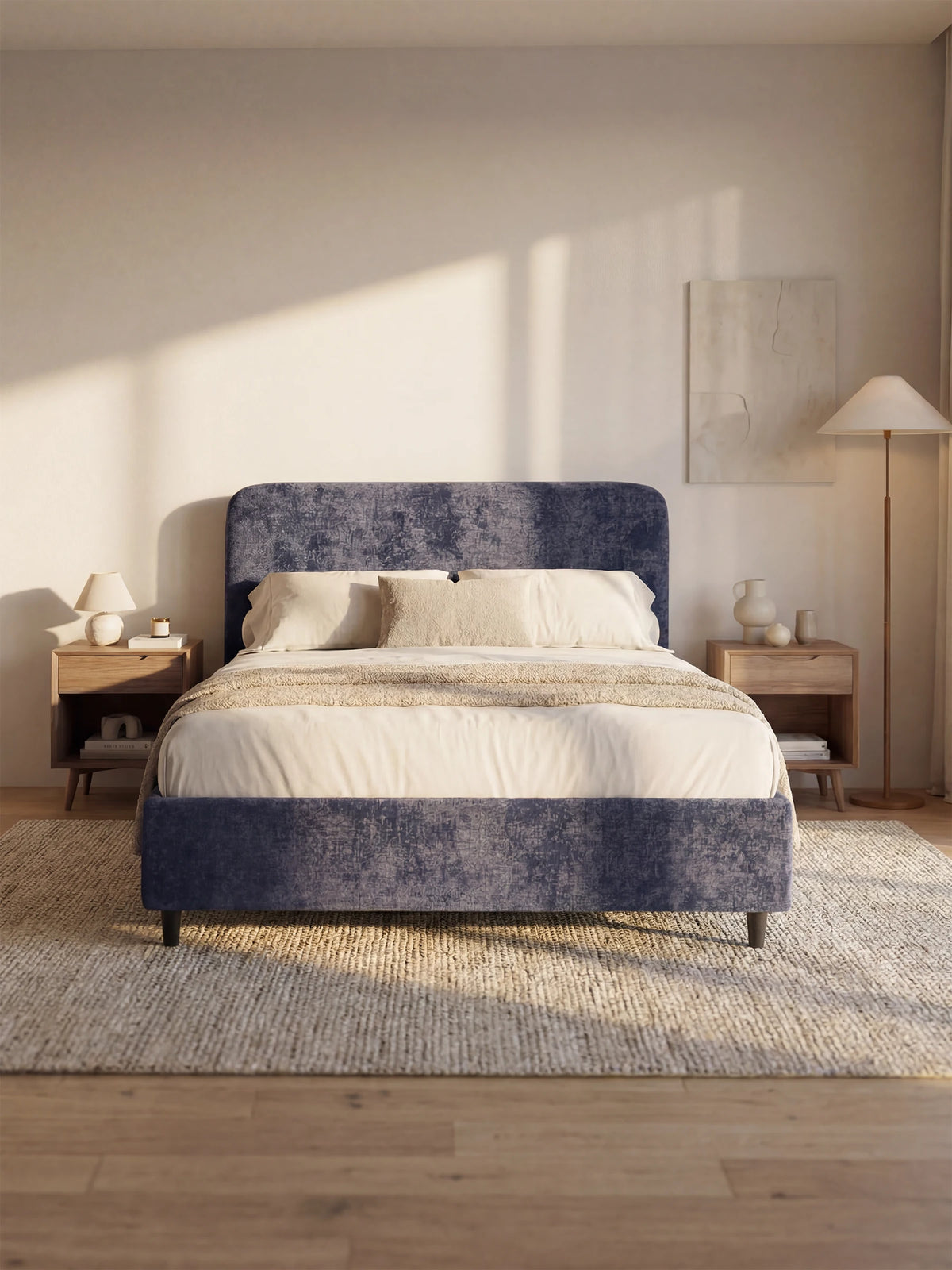 Un letto KAELOS minimal imbottito, con una testiera arrotondata dalla trama blu-violetta, è collocato in una camera da letto luminosa e moderna, al centro di un tappeto intrecciato, tra due comodini in legno su cui sono disposti lampade, vasi e libri.