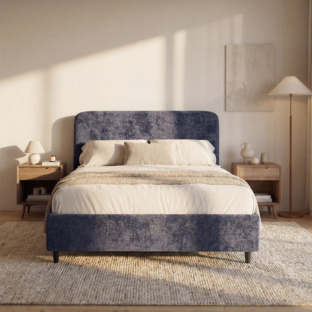 Un'accogliente camera da letto arredata con il letto KAELOS minimal imbottito di Letti.store: in ciniglia blu navy, con gambe alte, testiera arrotondata e vano contenitore. Il tutto completato da biancheria da letto bianca e beige, comodini in legno con lampade, quadri alle pareti e un tappeto beige dalla trama ricca, illuminato dalla calda luce del sole.