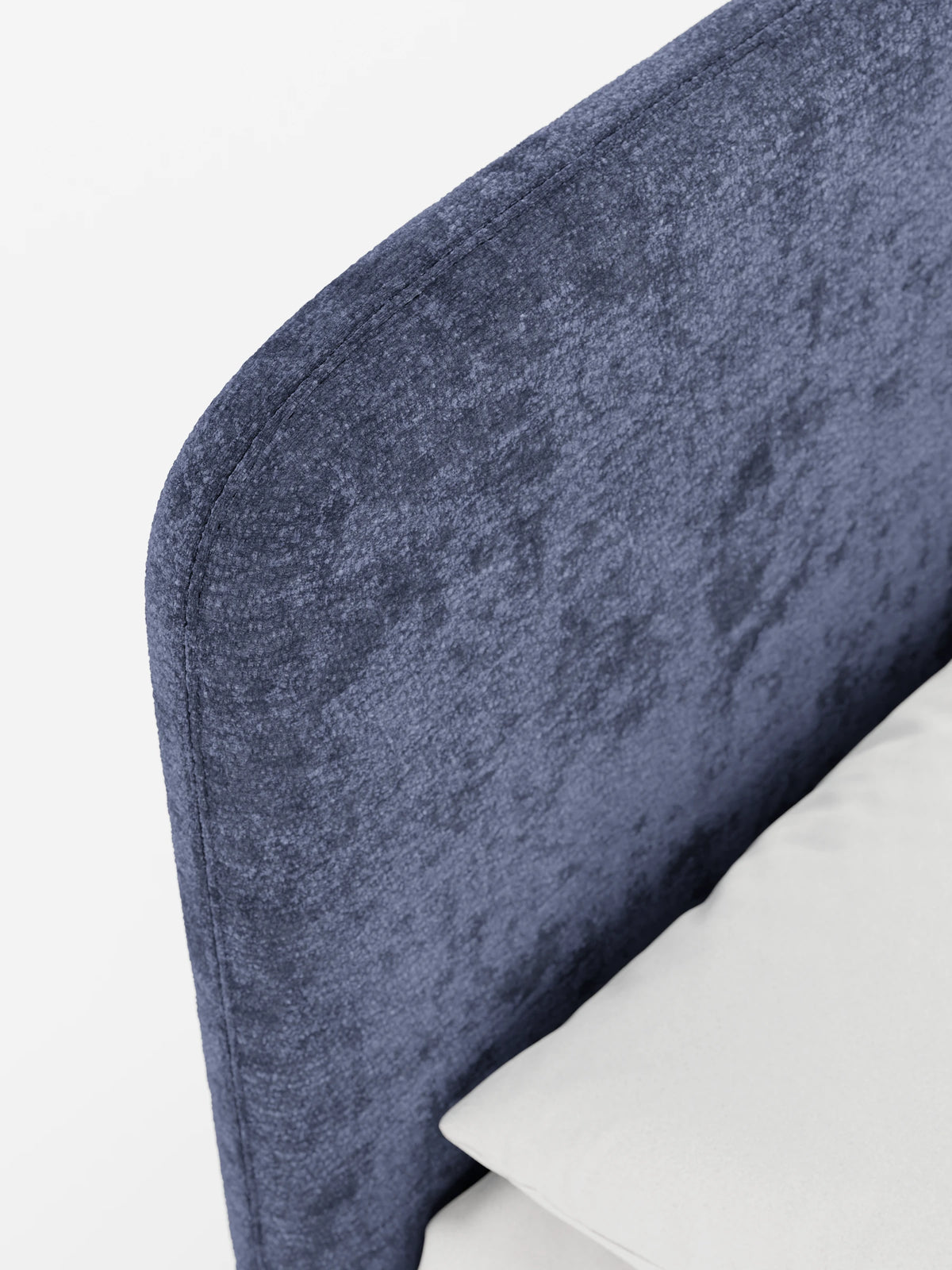 Un primo piano della testiera arrotondata in ciniglia blu di un letto KAELOS minimal imbottito - con contenitore, piedini alti e testiera arrotondata in tessuto ciniglia, su cui poggia un cuscino bianco contro il tessuto blu multitonale dalla trama rigata.