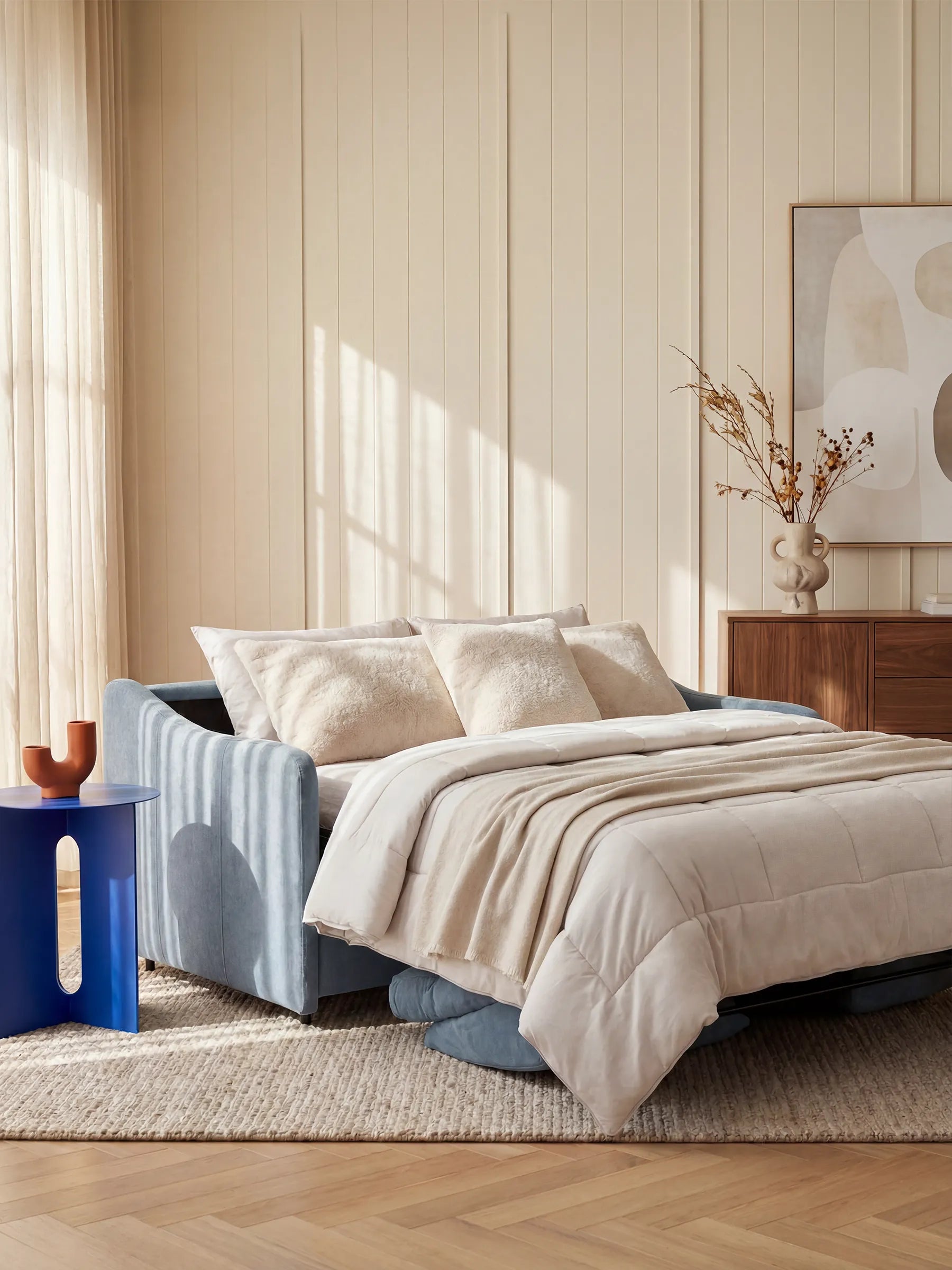 Una camera da letto accogliente è arredata con il divano letto Lodovico a 3 posti con materasso ALTO 20 cm in tessuto smacchiabile certificato Oeko-Tex di Divani.Store, in azzurro, con biancheria da letto e cuscini beige, posizionato su un tappeto intrecciato. Un tavolino blu con elementi decorativi, una cassettiera in legno, un quadro astratto e un vaso con rami secchi completano l’ambiente moderno e illuminato da luci soffuse.