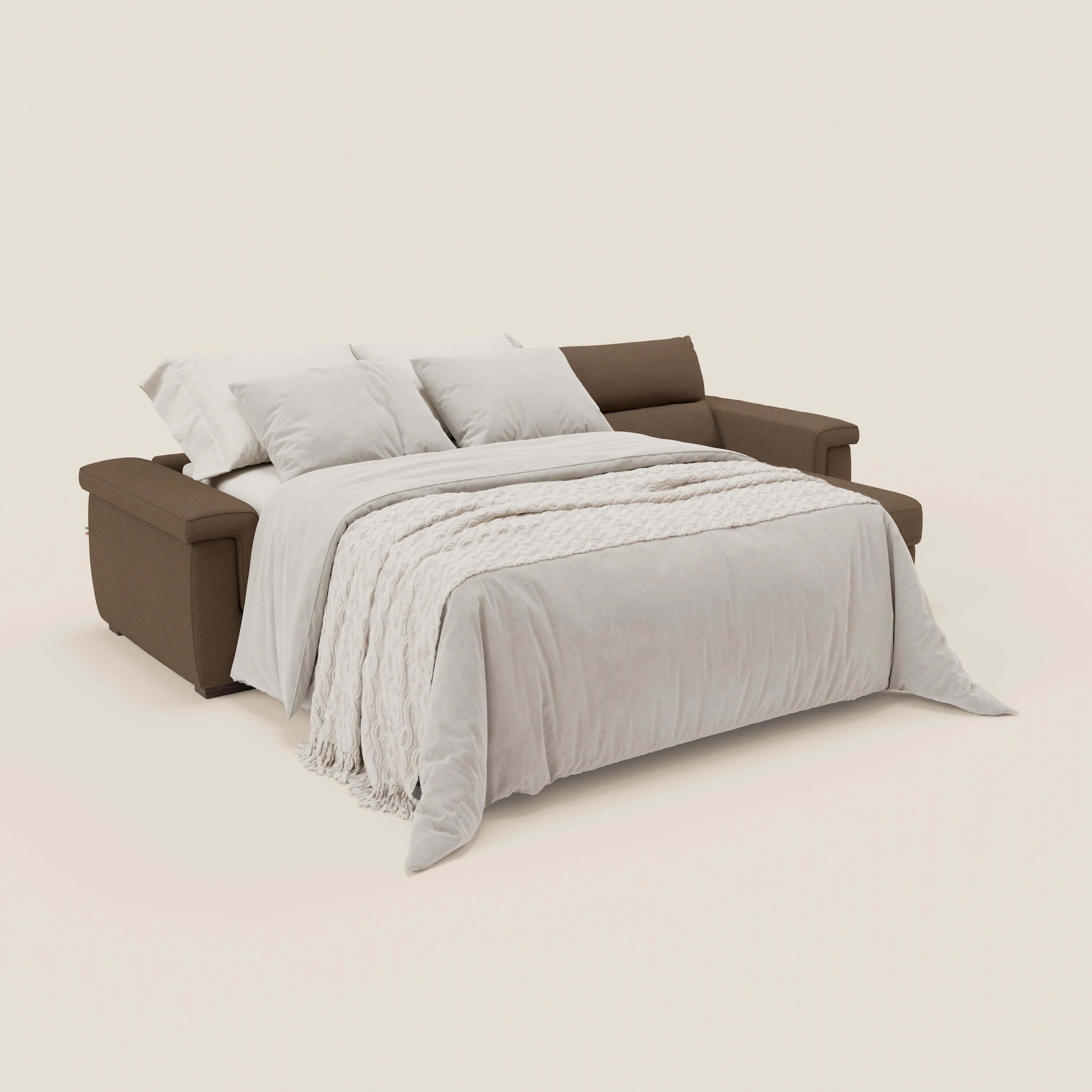 Il divano letto Magnolia angolare con penisola contenitore di Divani.Store in tessuto marrone T05 è aperto a letto, completato da cuscini bianchi e una coperta morbida. Lo sfondo crema esalta la praticità nascosta di questo accogliente divano letto.