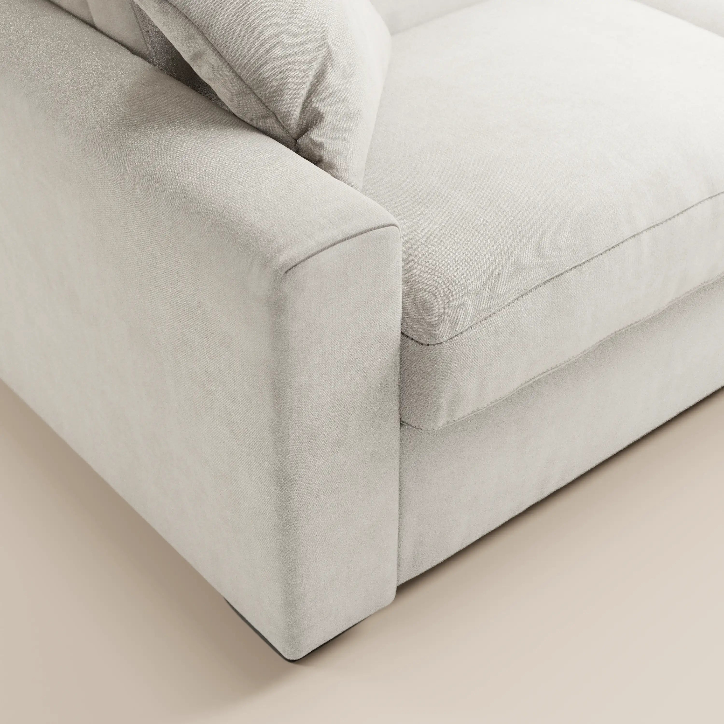 Primo piano di un divano letto Maximus a 3 posti con materasso ALTO 20 cm di Divani.Store, dotato di poggiatesta regolabili e rivestito in tessuto Oeko-Tex antimacchia color beige chiaro; nell'immagine sono visibili il bracciolo, la seduta, il cuscino morbido e una parte del pavimento chiaro.