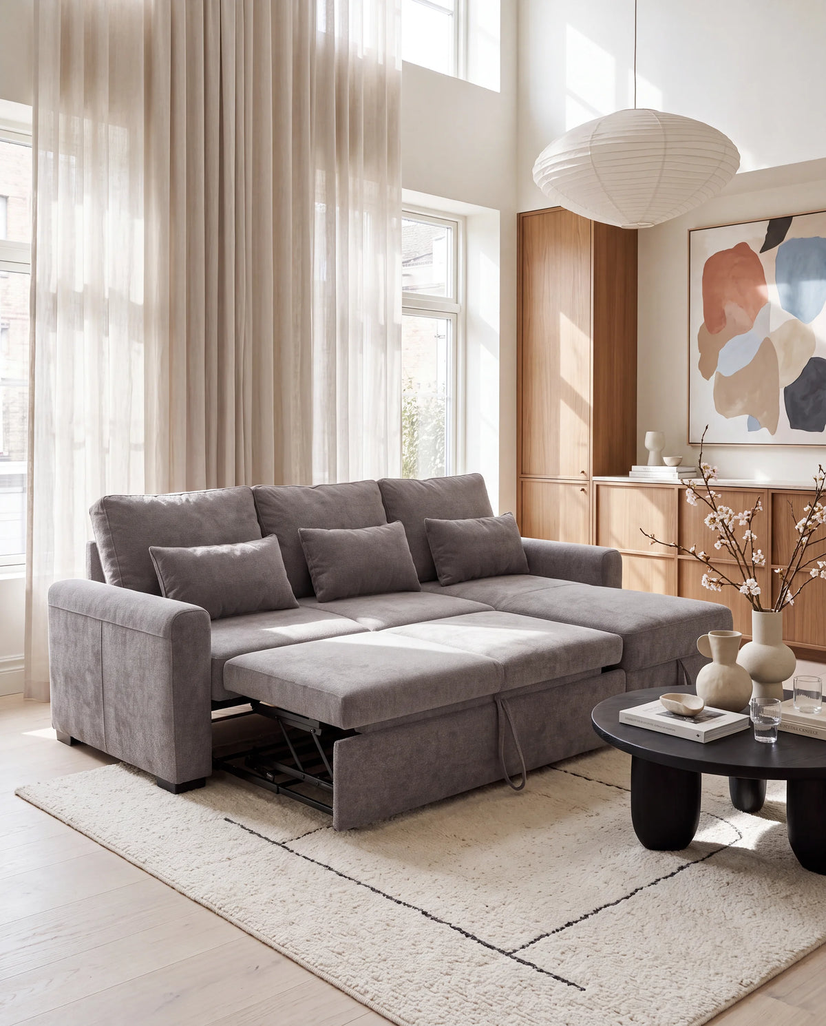 Un divano letto Ottaviano con letto estraibile, penisola reversibile e contenitore in tessuto materico smacchiabile è collocato in un luminoso e moderno soggiorno su un tappeto color crema accanto a un tavolino da caffè, con tende trasparenti e mobili in legno sullo sfondo.