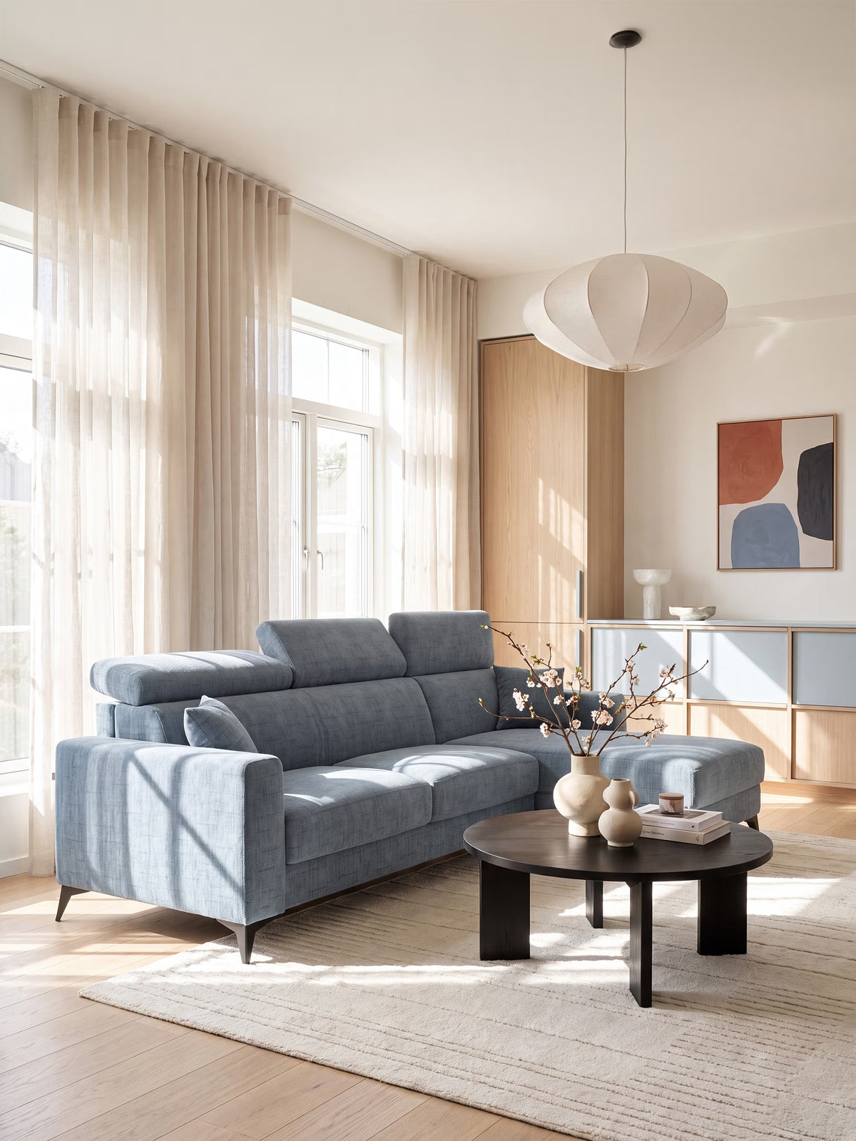 Un soggiorno moderno arredato con il divano letto angolare Pegaso di Divani.Store, dotato di poggiatesta regolabili e di una chaise longue con vano contenitore, rivestito in tessuto jacquard T22 antimacchia di colore azzurro. Lo spazio comprende un tavolino rotondo color caffè scuro impreziosito da vasi e rami color beige, pavimenti in legno chiaro, un tappeto dai toni neutri, opere d’arte astratte alle pareti, una lampada a sospensione, ampie finestre adornate da tende trasparenti e mobili su misura in