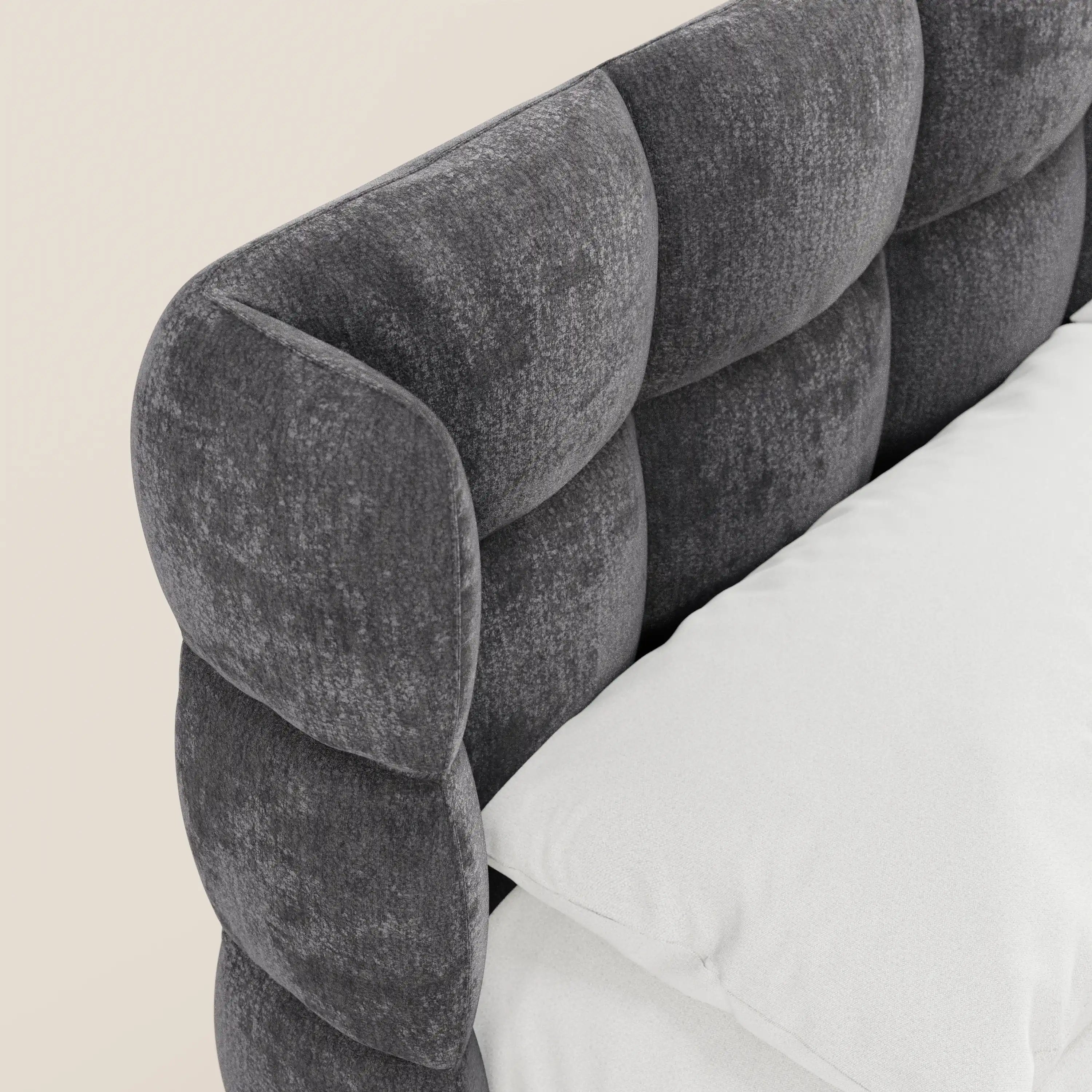 Primo piano del letto imbottito RAGNAR con contenitore e testiera capitonné avvolgente di Letti.store, realizzato in un elegante tessuto grigio scuro certificato Oeko-Tex® con motivo a griglia. Un cuscino e un materasso bianchi risaltano su uno sfondo beige chiaro.