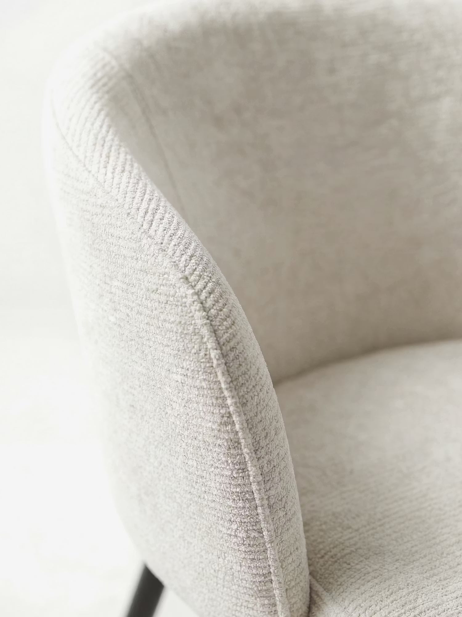 Primo piano della sedia Rovo con braccioli di Divani.Store: una sedia moderna con schienale ricurvo in morbida ciniglia beige chiaro e gambe in metallo nero parzialmente visibili. Vengono messi in risalto la consistenza soffice e i contorni delicati.