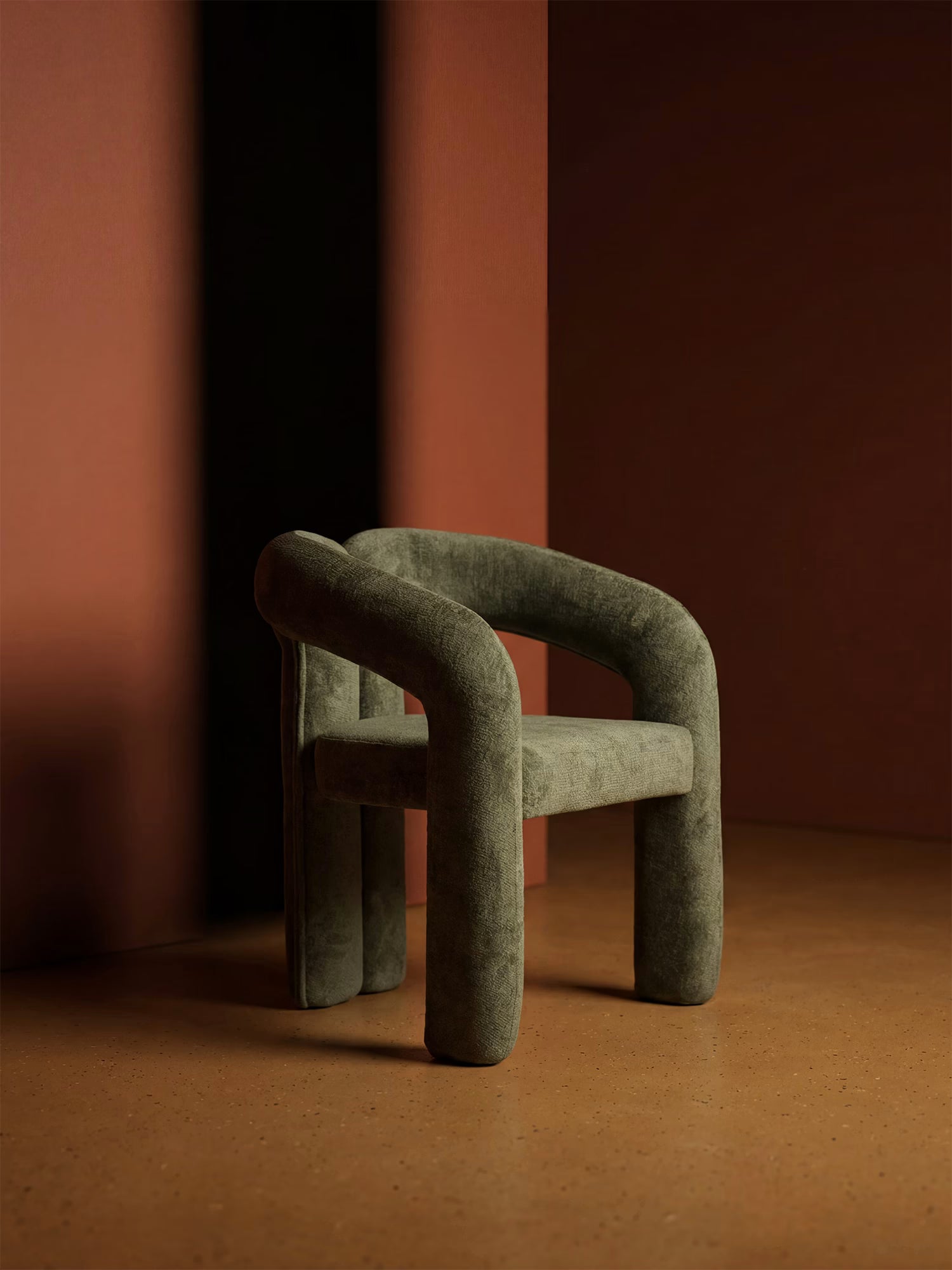 La sedia Sibilla di Divani.Store, imbottita e rivestita in tessuto con struttura ad arco, è collocata su un pavimento marrone a pois davanti a pareti scure e color ruggine, creando un ambiente caldo, minimalista e contemporaneo.