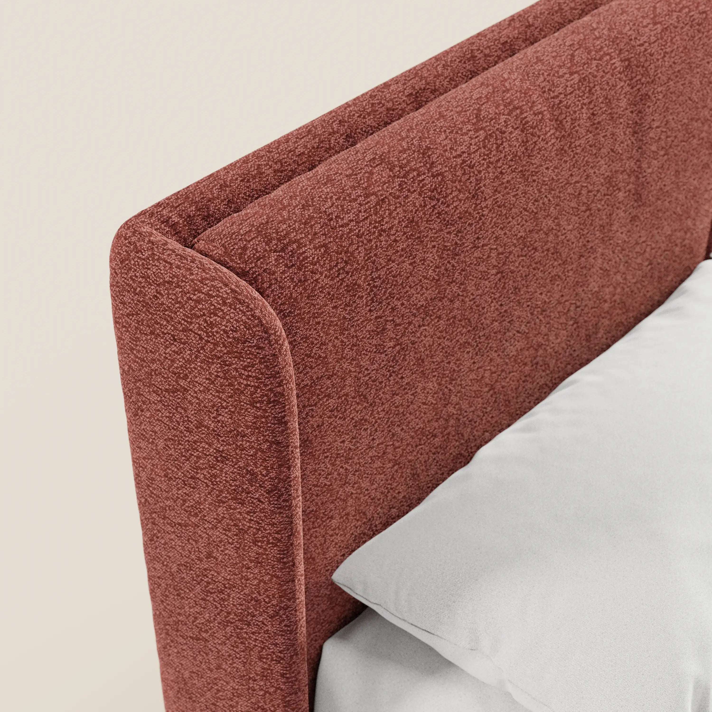 Primo piano del letto di design VALKIRIA di Letti.store, caratterizzato da una testiera avvolgente rivestita in tessuto Oeko-Tex® marrone-rossastro a trama ruvida con 2 cuscini imbottiti. Sullo sfondo beige chiaro si notano un cuscino bianco e un lenzuolo.