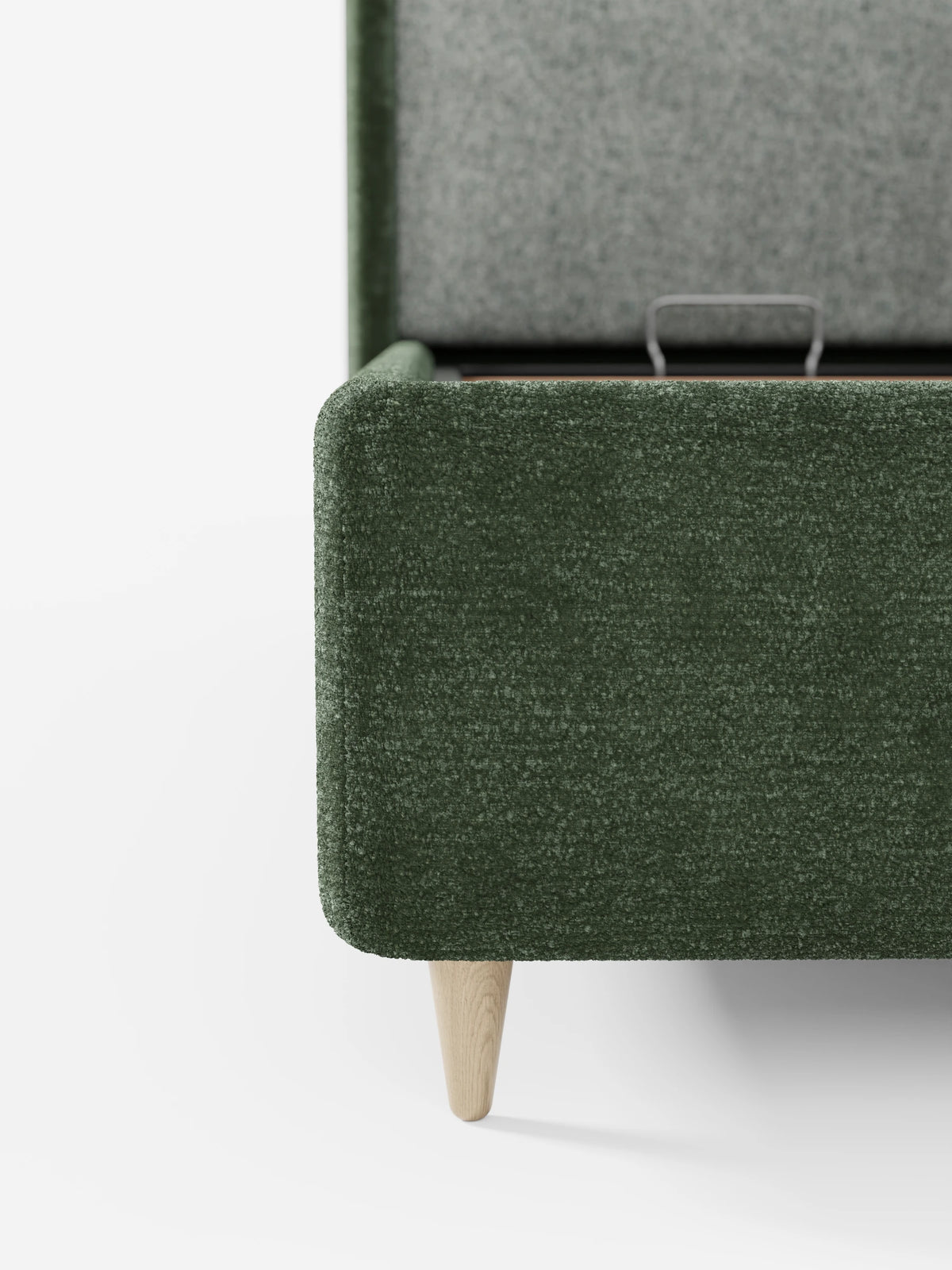 Un primo piano di un letto imbottito bicolore VALKIRIA color verde bosco con contenitore, con lo scomparto parzialmente aperto; il letto presenta un angolo arrotondato su una gamba in legno chiaro su sfondo bianco, da cui spunta una maniglia metallica.