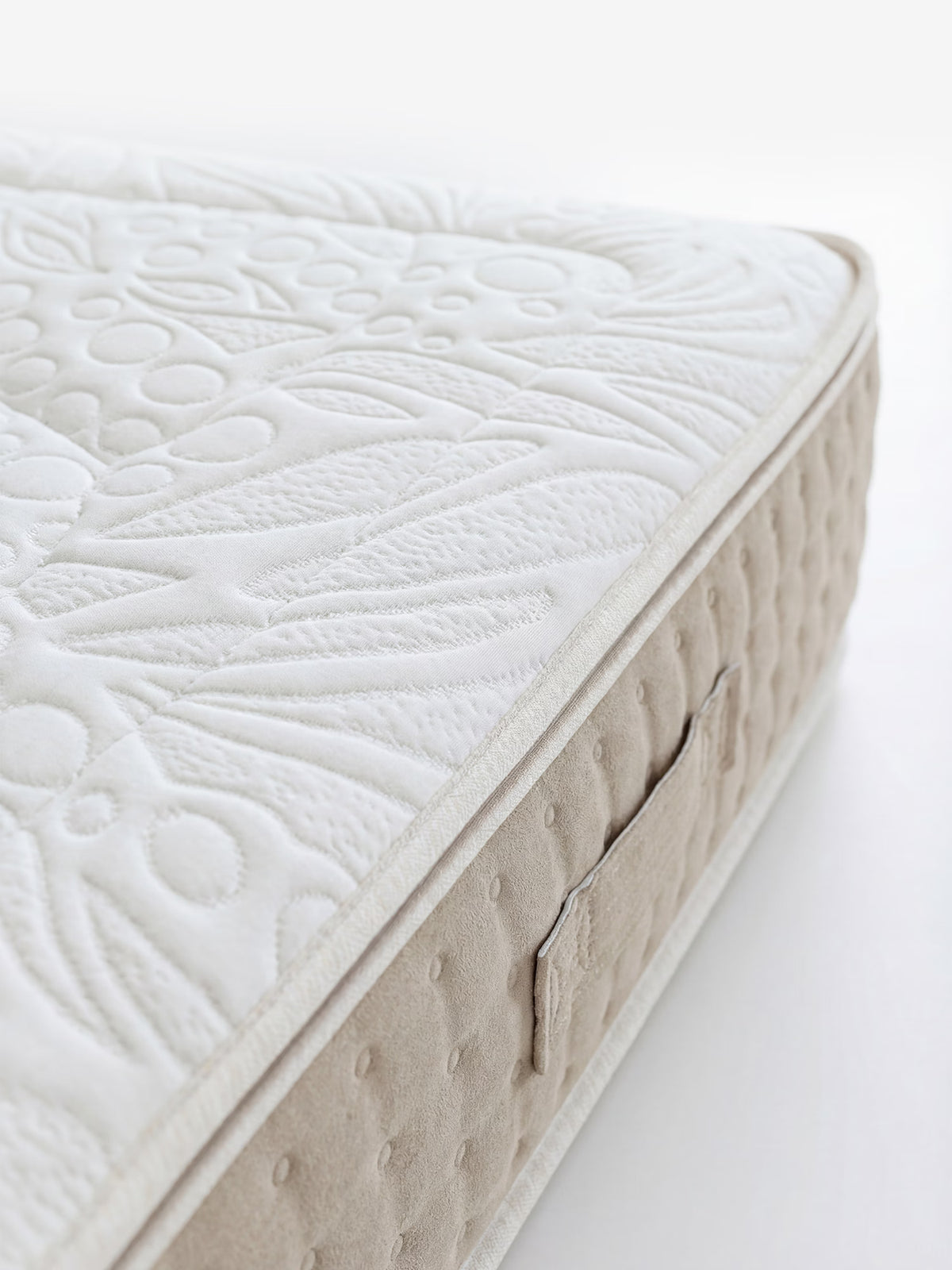 Un primo piano del modello VELTHA – Materasso Bio-Adattivo con molle insacchettate e lattice naturale a 7 zone di Divani.Store mostra un tessuto bianco testurizzato con motivi ricamati e un pannello laterale beige, caratterizzato da piccoli puntini in rilievo e da una maniglia in tessuto cucita. Lo sfondo semplice e luminoso mette in risalto la lavorazione artigianale curata nei minimi dettagli del materasso.