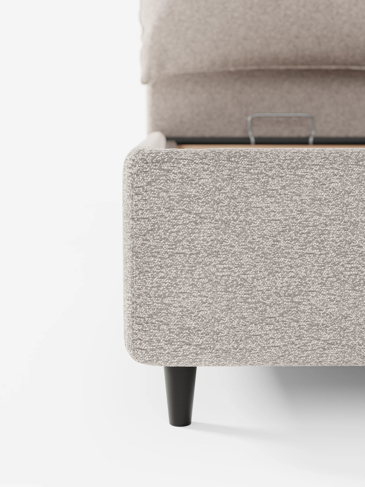 Un primo piano del letto imbottito ZAYRON con contenitore e morbidi cuscini regolabili, caratterizzato da un angolo rivestito in tessuto grigio chiaro con effetto testurizzato, una maniglia in metallo per l’apertura del contenitore e una gamba corta e conica di colore scuro.