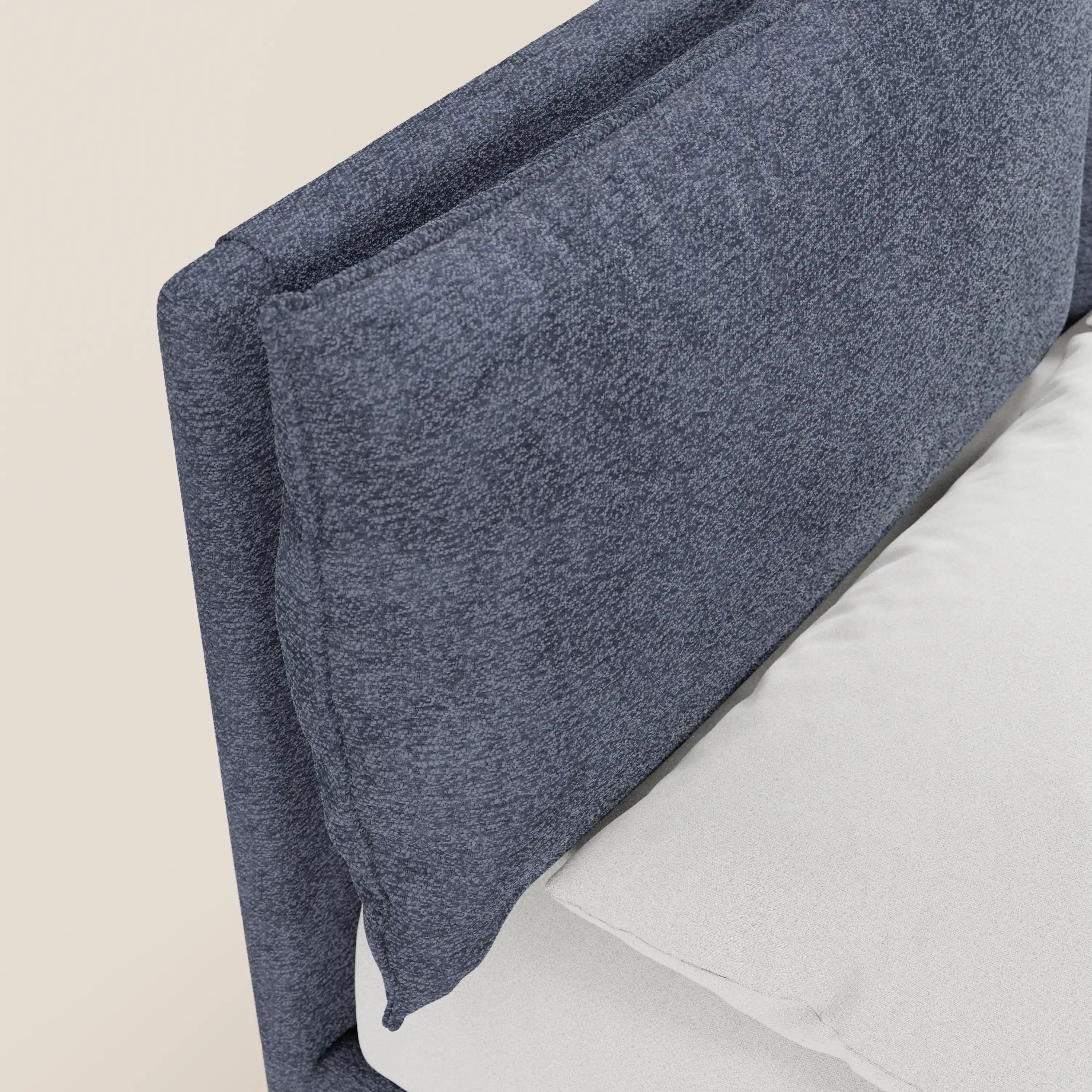 Primo piano del letto imbottito ZAYRON di Letti.store, caratterizzato da una testiera in tessuto blu scuro certificato Oeko-Tex® con cuscini regolabili e materasso bianco; lo sfondo è di colore beige chiaro a tinta unita.