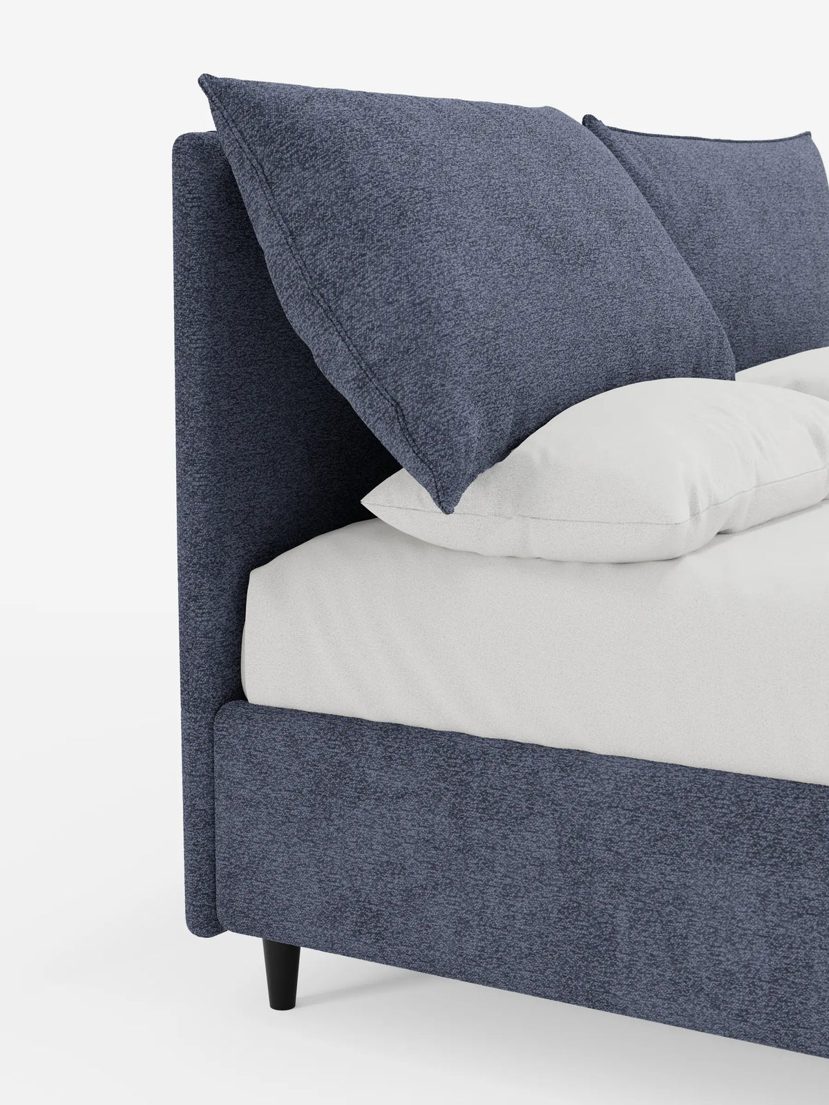 Primo piano di un letto imbottito ZAYRON con contenitore e morbidi cuscini regolabili in tessuto blu scuro testurizzato, caratterizzato da grandi cuscini angolari disposti sulla biancheria da letto bianca su uno sfondo bianco a tinta unita.