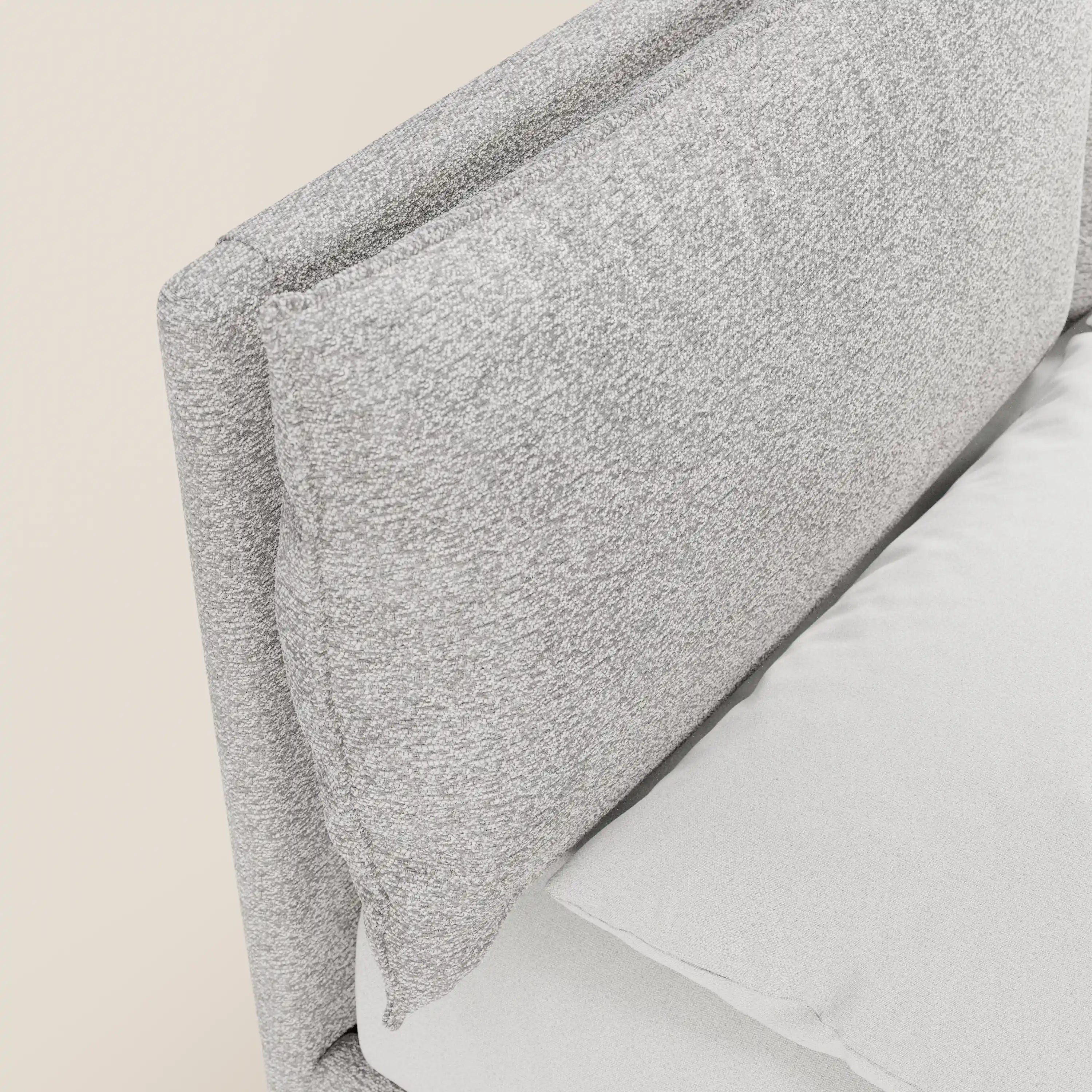 Primo piano del letto imbottito ZAYRON di Letti.store, caratterizzato da una testiera in tessuto grigio chiaro certificato Oeko-Tex® e da cuscini regolabili. Un cuscino coordinato completa il design, con un lenzuolo bianco e uno sfondo beige a tinta unita.