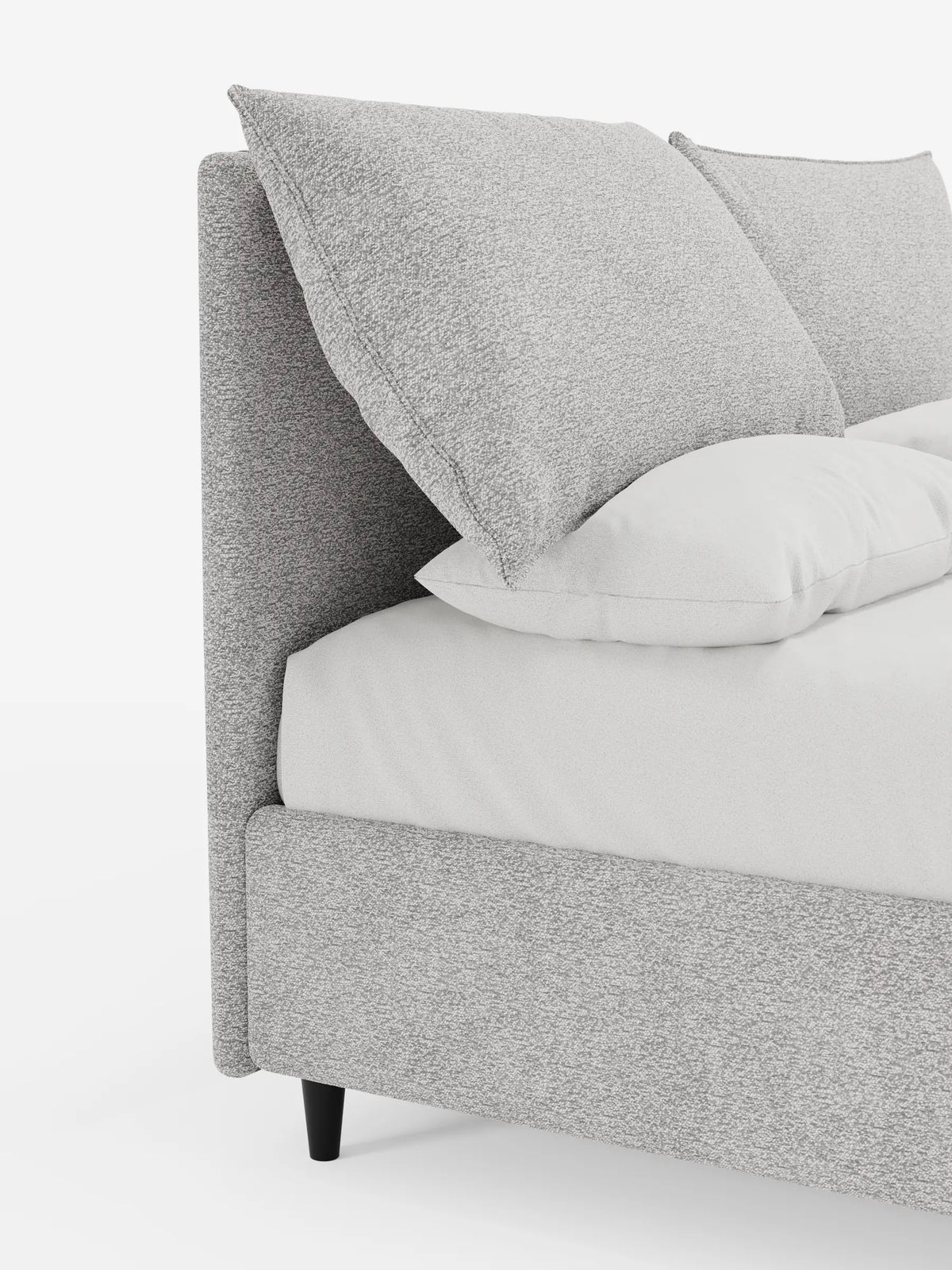 Primo piano di un letto imbottito ZAYRON con contenitore e morbidi cuscini regolabili, caratterizzato da una struttura rivestita in grigio chiaro con due grandi cuscini rettangolari inclinati sopra un lenzuolo bianco, appoggiato su piccole gambe scure.