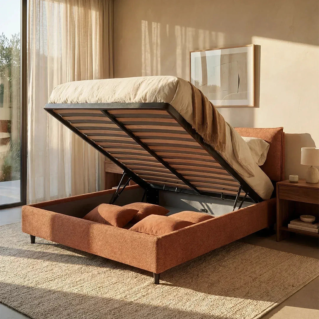 In una camera da letto inondata di luce solare spicca il letto imbottito con contenitore ZAYRON di Letti.store, con il materasso sollevato per rivelare lo spazio dedicato ai cuscini. L'ambiente è completato da un tappeto beige, un comodino in legno, tende trasparenti e un quadro incorniciato appeso alla parete sopra il letto.