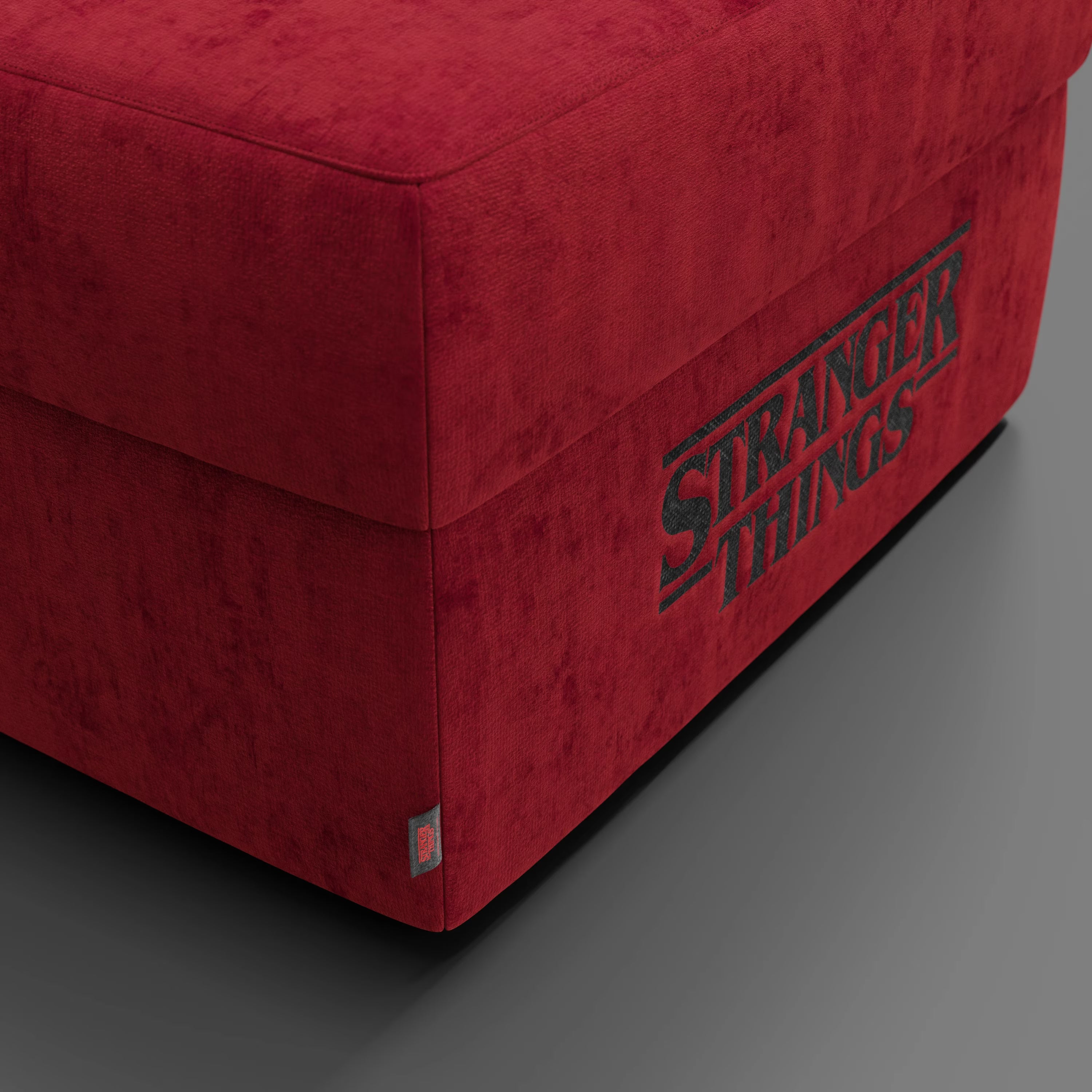 Il pouf Divani.Store Sun Contenitore Stranger Things Edition (60x80 cm, rosso, tessuto smacchiabile di Netflix) presenta il logo nero della serie ricamato sul lato e una piccola etichetta in tessuto nell'angolo inferiore.