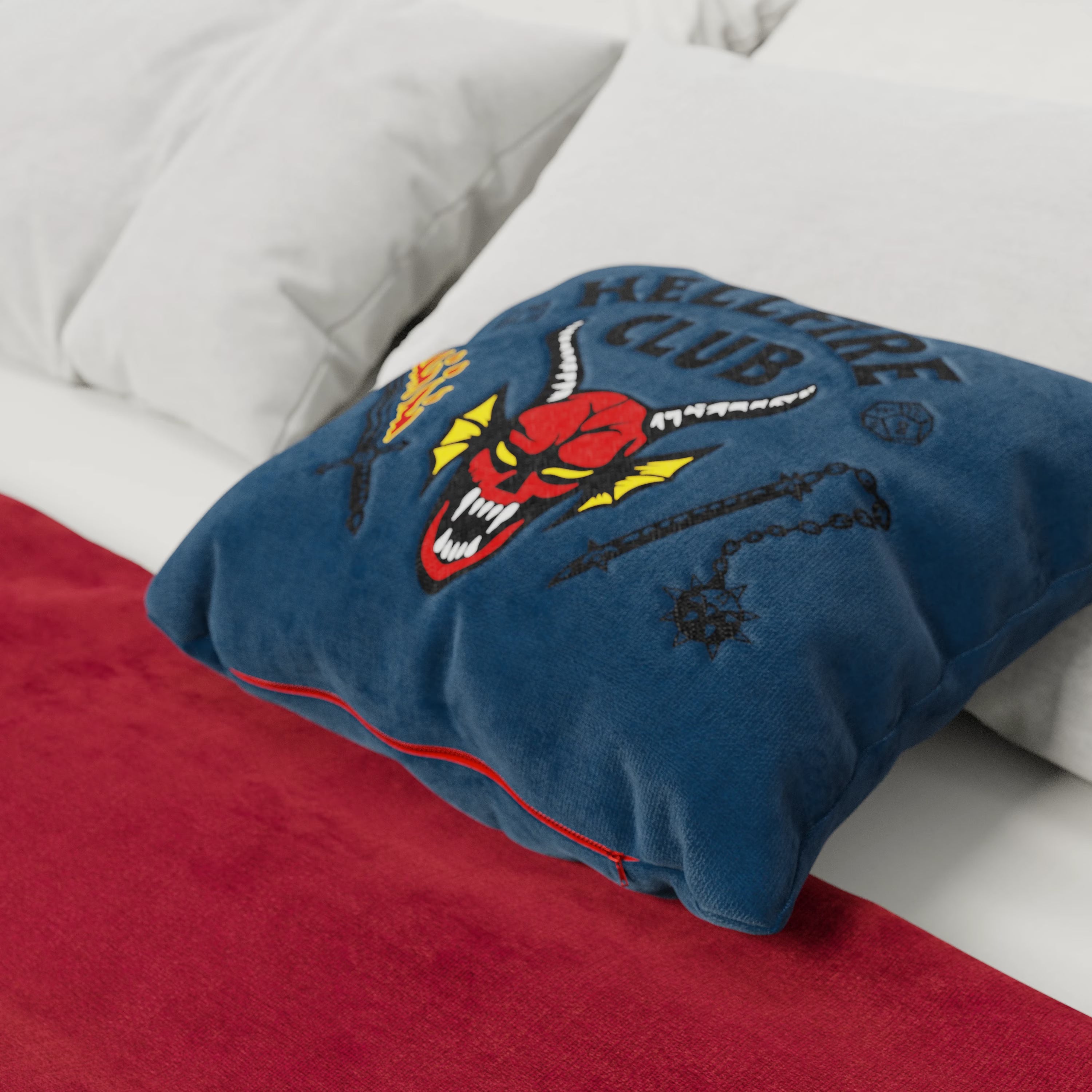 Un Cuscino Hellfire club Stranger Things Limited Edition di Divani.Store, 45x45 cm, smacchiabile in tessuto blu con ricami audaci, tra cui la faccia di un demone e il testo "HELLFIRE CLUB", poggia su un letto bianco. Presenta una cerniera rossa, perfetta per i fan!.