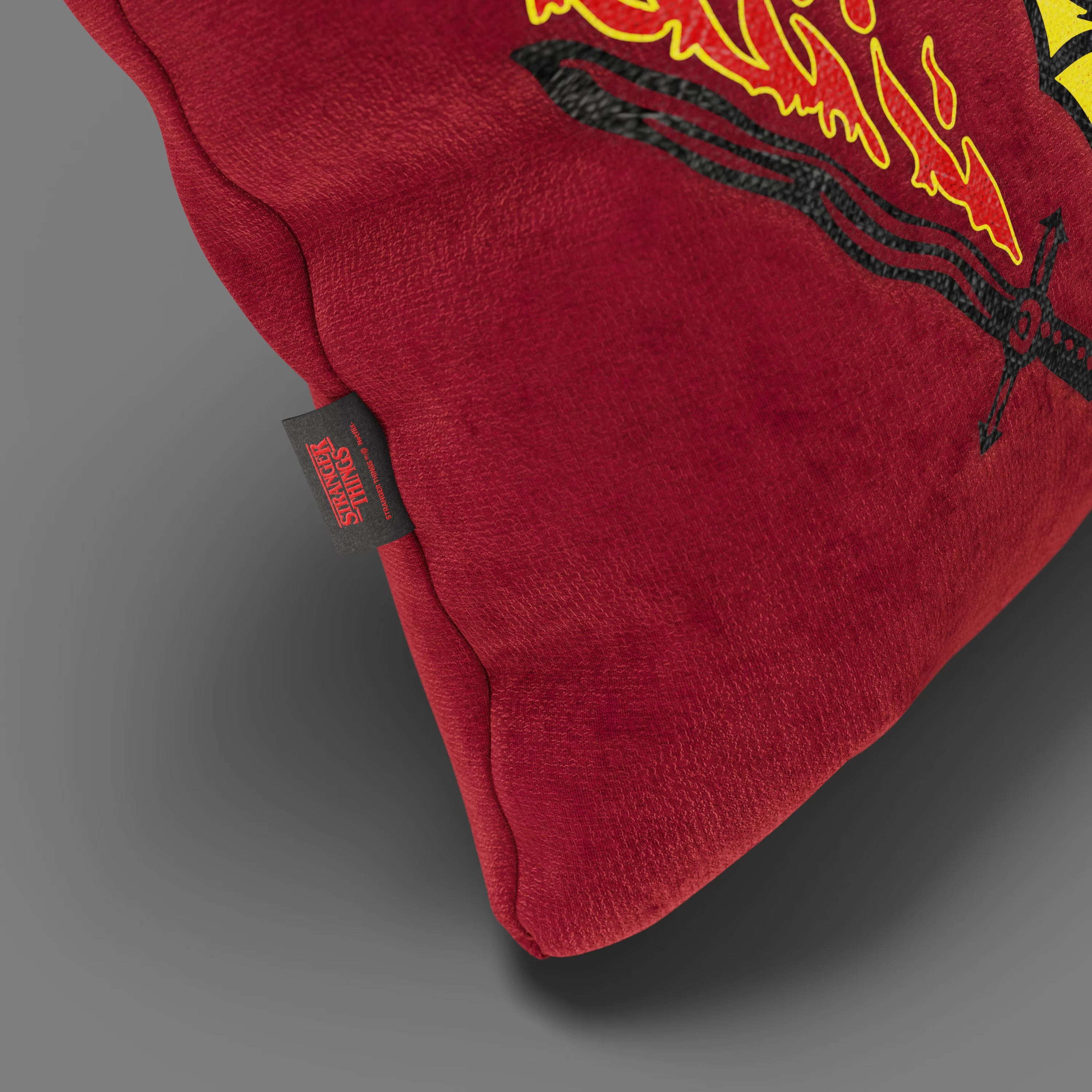Primo piano del cuscino Divani.Store Hellfire Club Stranger Things Limited Edition (45x45 cm) in rosso, con fiamme ricamate e una spada nera, con etichetta "Stranger Things" su tessuto smacchiabile, su sfondo grigio.